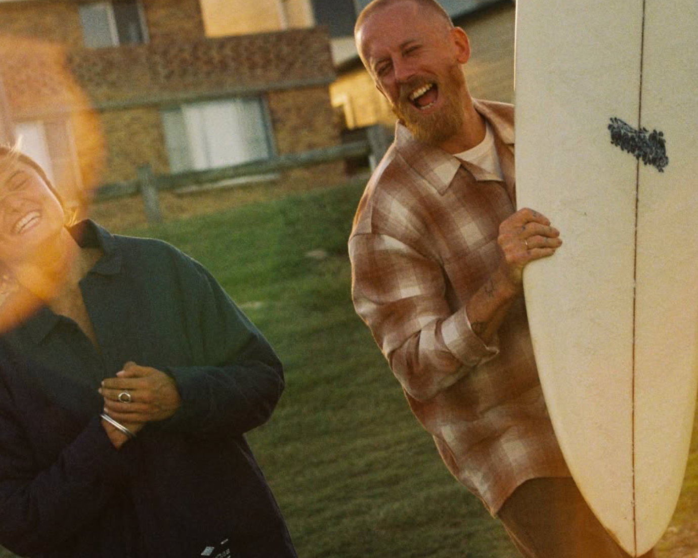 Two people laughing outdoors at sunset, one man holding a surfboard, wearing a plaid shirt, and the other person in a dark jacket with their hands on their chest.
