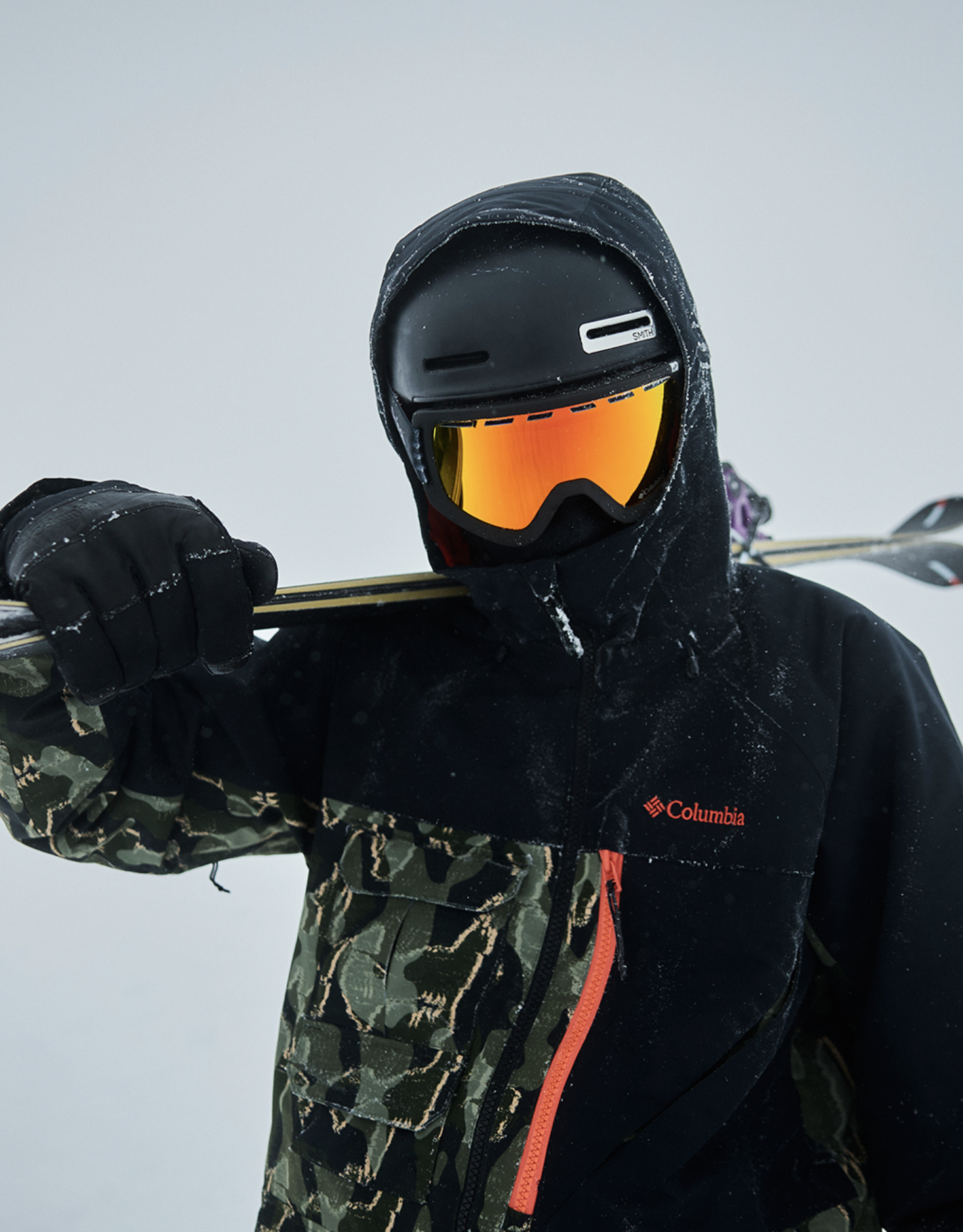 A person dressed for skiing, wearing a black jacket with a Columbia logo, camouflage-patterned pants, black gloves, a black helmet, and orange-tinted ski goggles, holding skis over their shoulder.