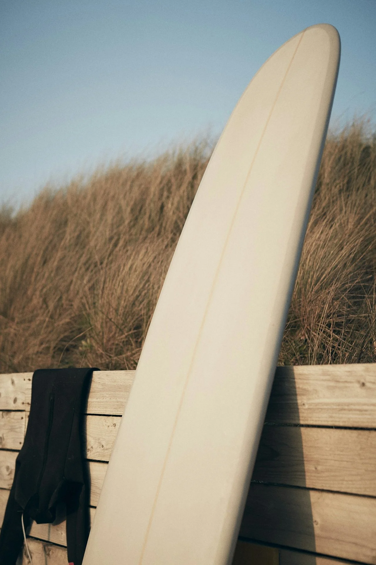 Close-up of a white surfboard leaning against a wooden fence on a beach, with sand dunes and a clear blue sky in the background.