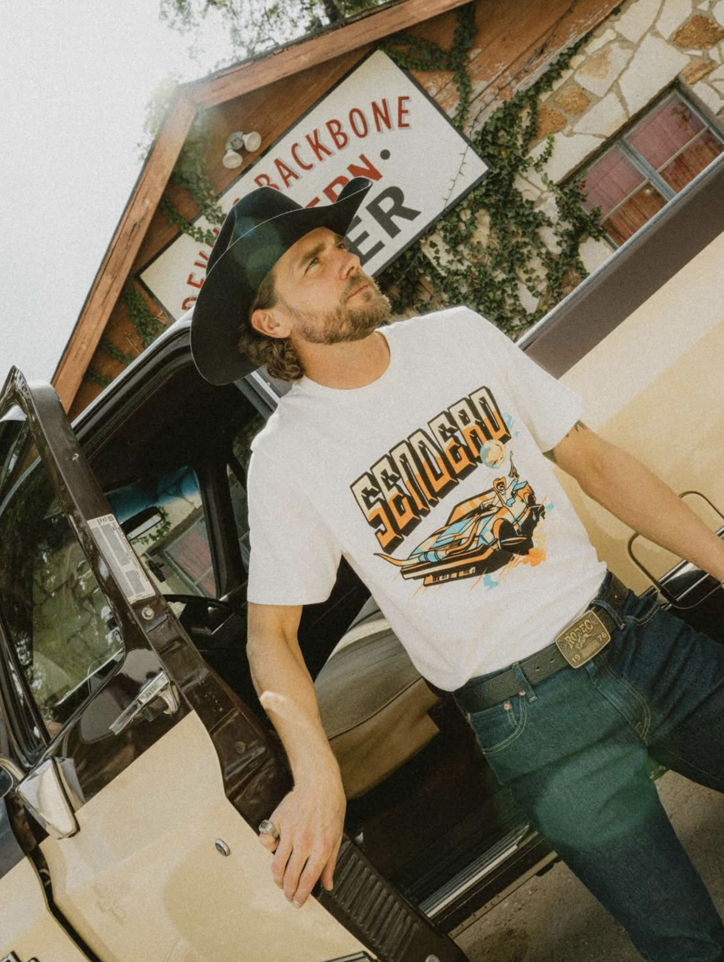 A man with a beard and long hair, wearing a cowboy hat and a graphic t-shirt, is standing next to a vintage car in a showroom. He has a belt with a large buckle and a ring on his right hand. Behind him, there is a building with a brick wall, a window, and a sign that reads 'Backbone.'