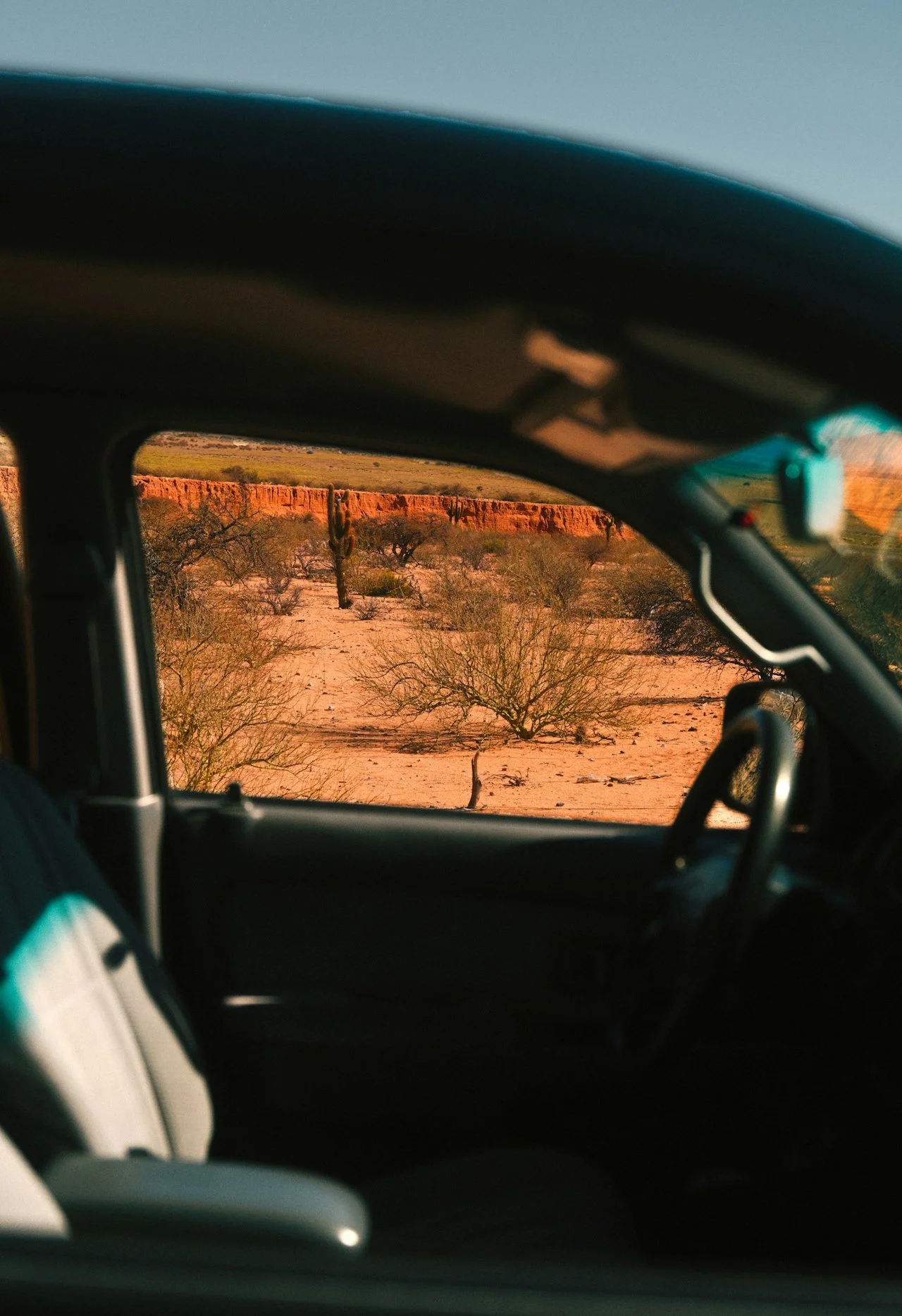 View through a vehicle's windshield of a desert landscape with scrubby bushes, cacti, and a reddish cliff in the distance at sunset or sunrise.