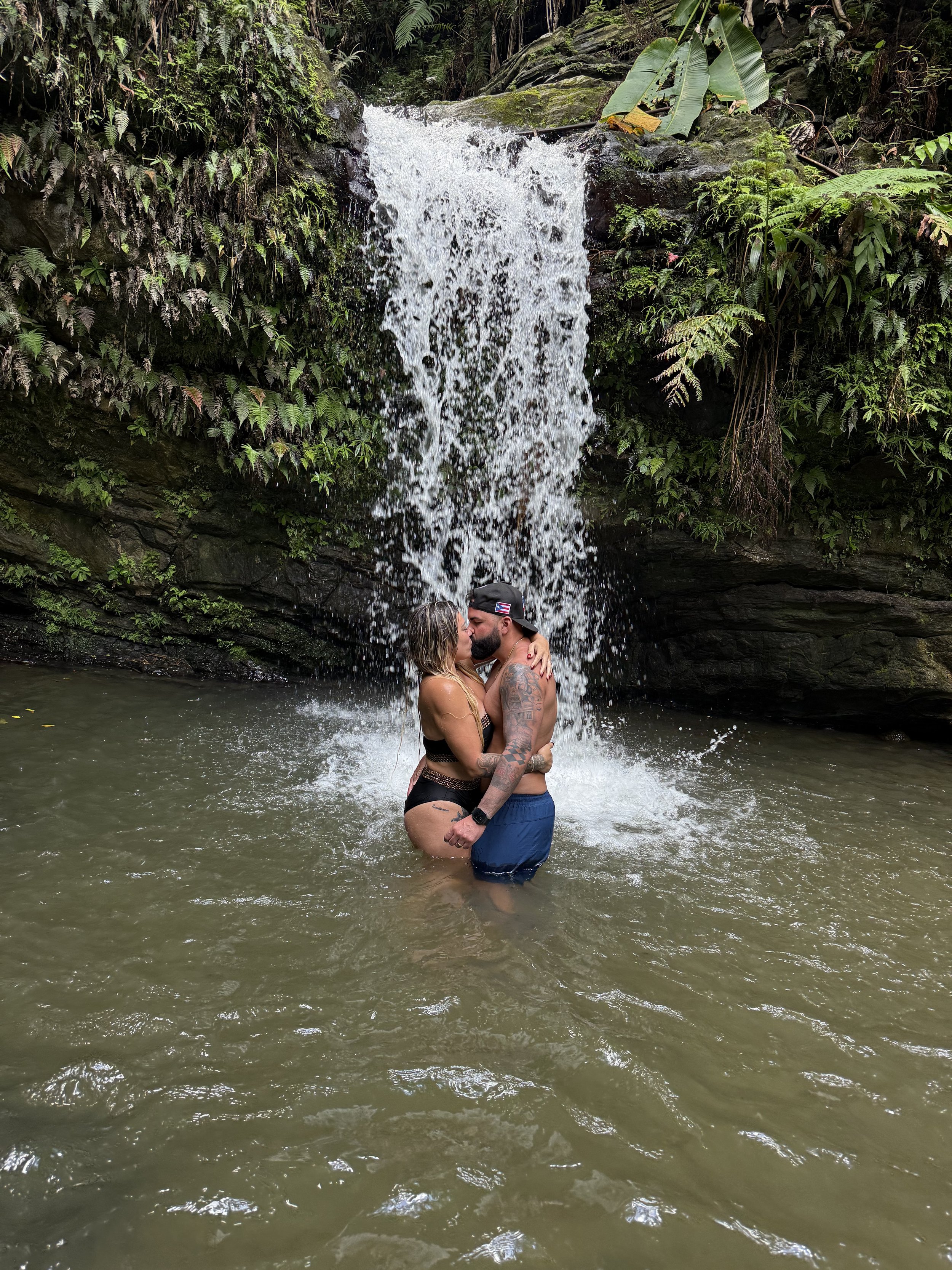 Pareja besándose frente a una cascada rodeada de naturaleza.
