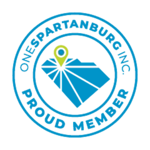 A blue circular logo with the outline of South Carolina at the center, a location pin, and rays emanating from the pin. The text around the circle reads 'ONE SPARTANBURG INC. PROUD MEMBER'.