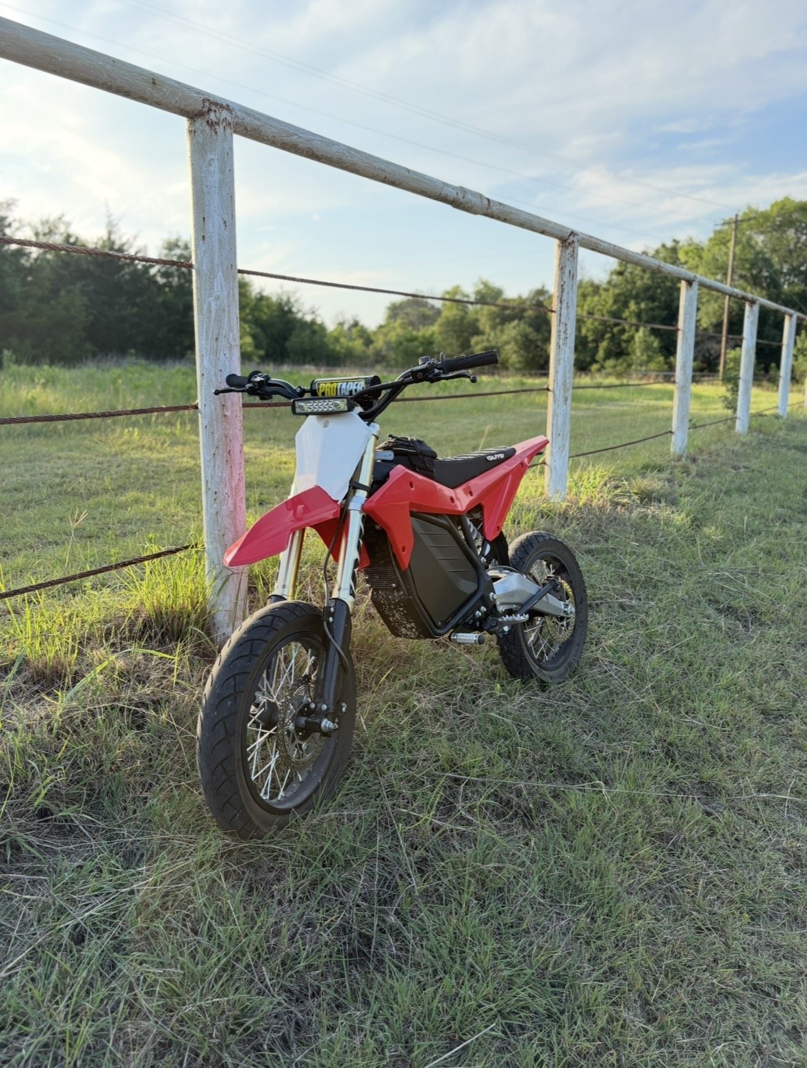 Electric dirt bike parked on grassy field next to a white wooden fence with a rural landscape in the background.