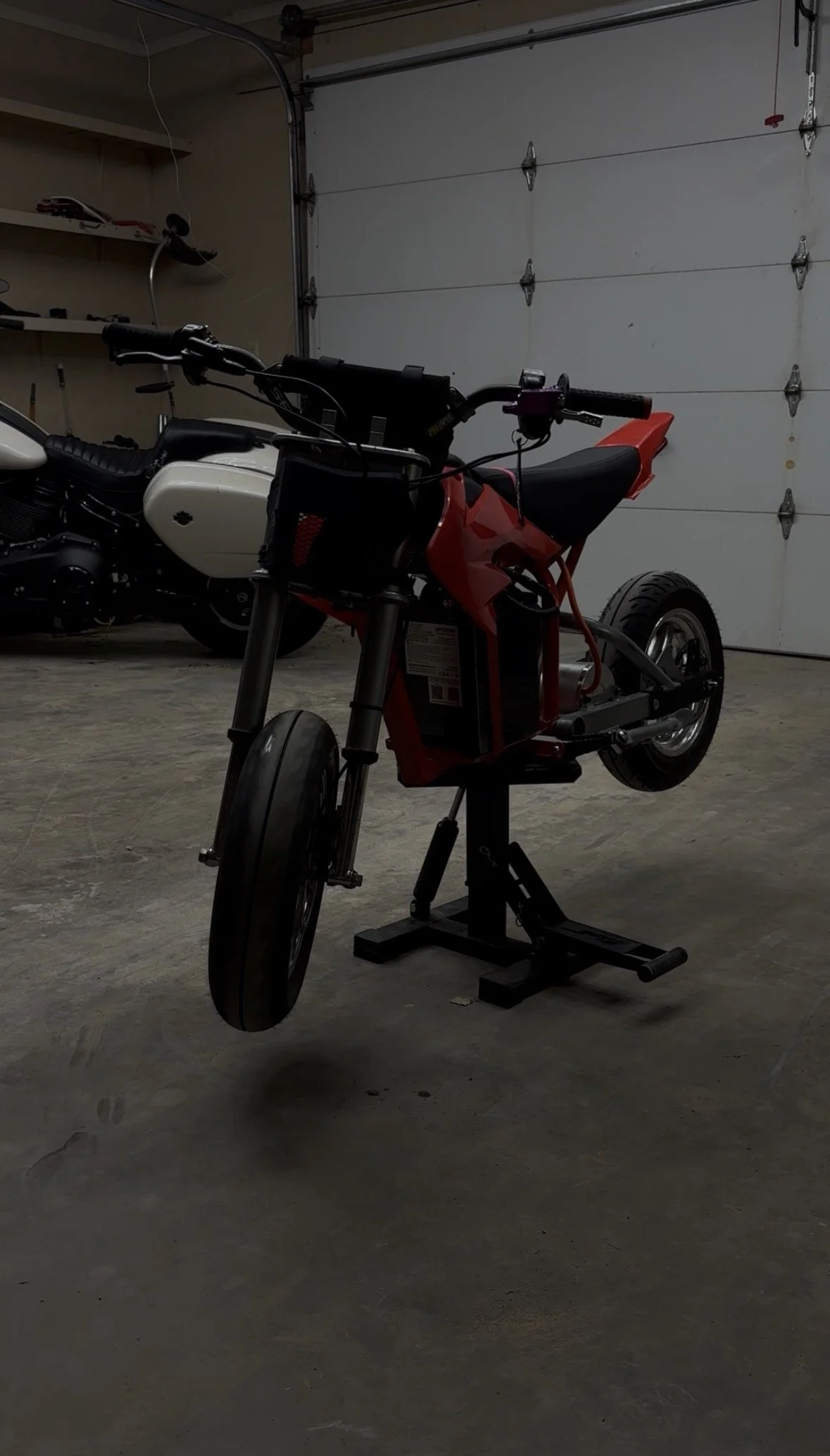 A red electric bicycle or motorcycle frame on a stand inside a garage next to a white motorcycle, with a garage door in the background.