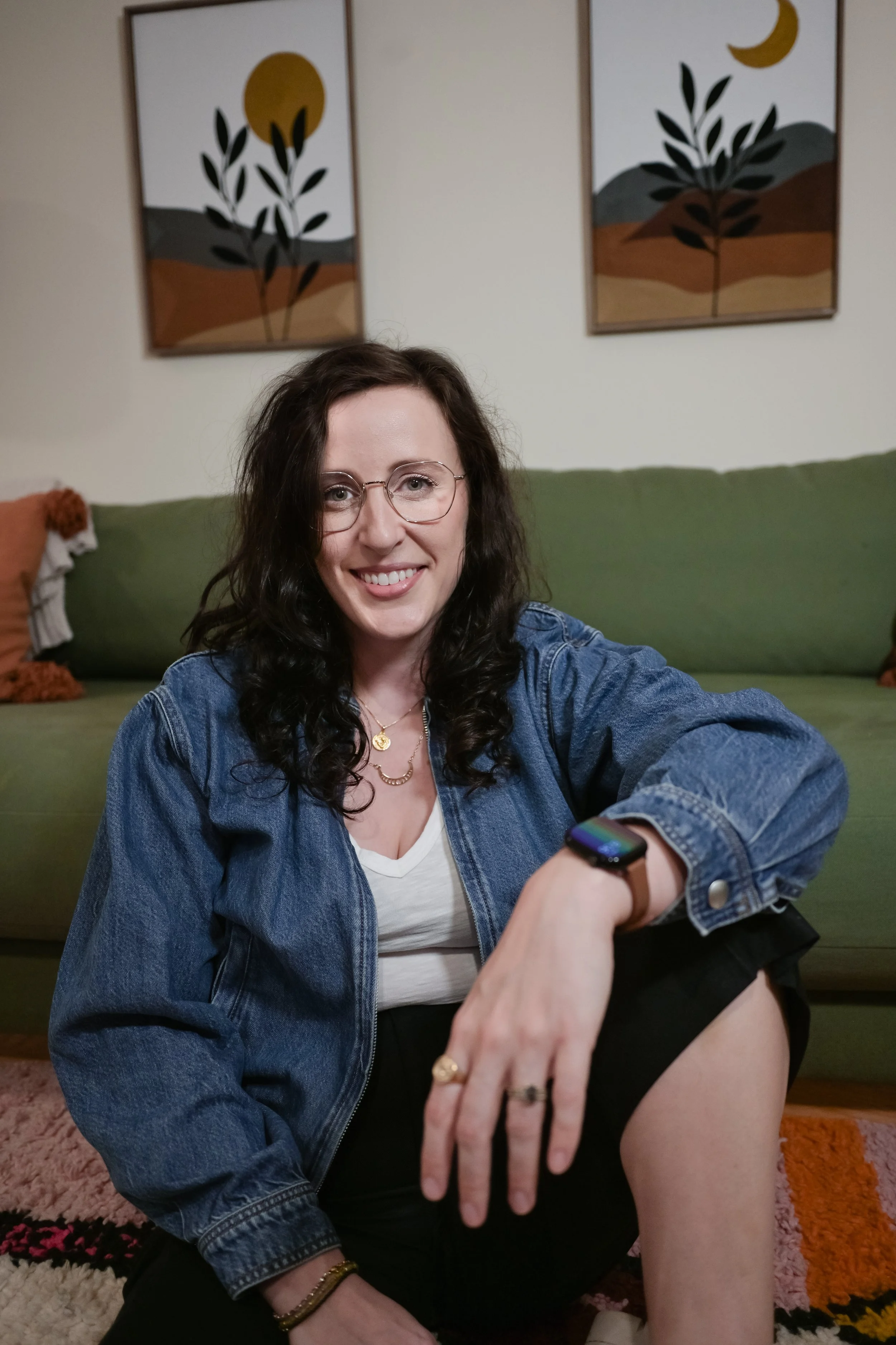 A woman with dark curly hair, glasses, and a denim jacket smiling while taking a selfie in a living room. The background features framed abstract landscape art on the wall and a green sofa with decorative pillows.