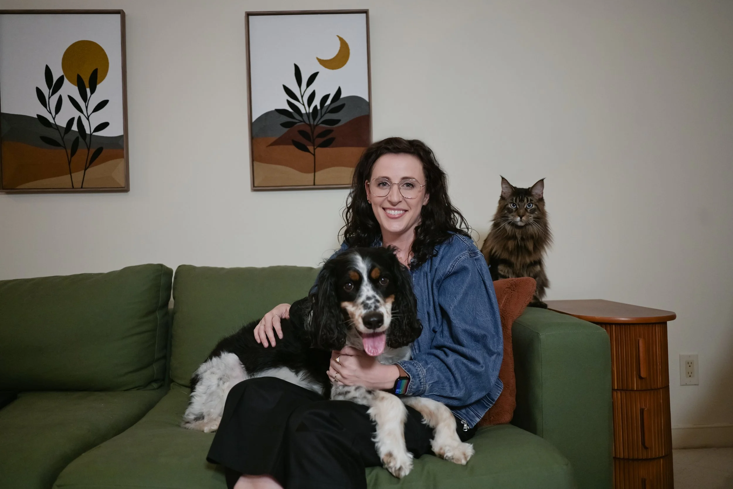 A woman with dark curly hair and glasses sitting on a green couch, smiling while holding a large black and white dog, with a tabby cat on a side table behind her. There are two abstract landscape paintings on the cream wall behind them.
