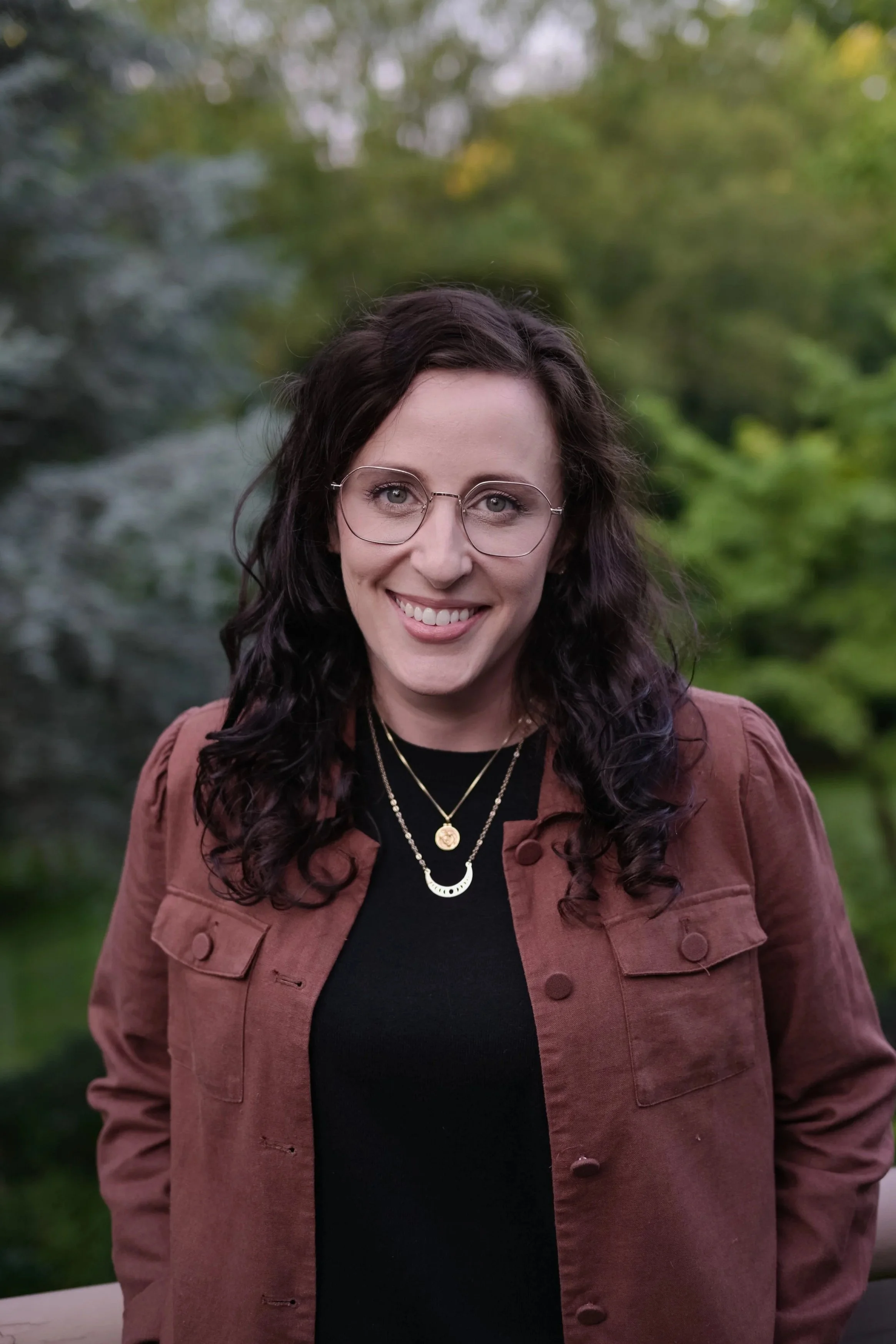 A woman with curly dark hair and glasses smiling outdoors, wearing a black top, layered necklaces, and a brown jacket, with a green, blurred natural background.