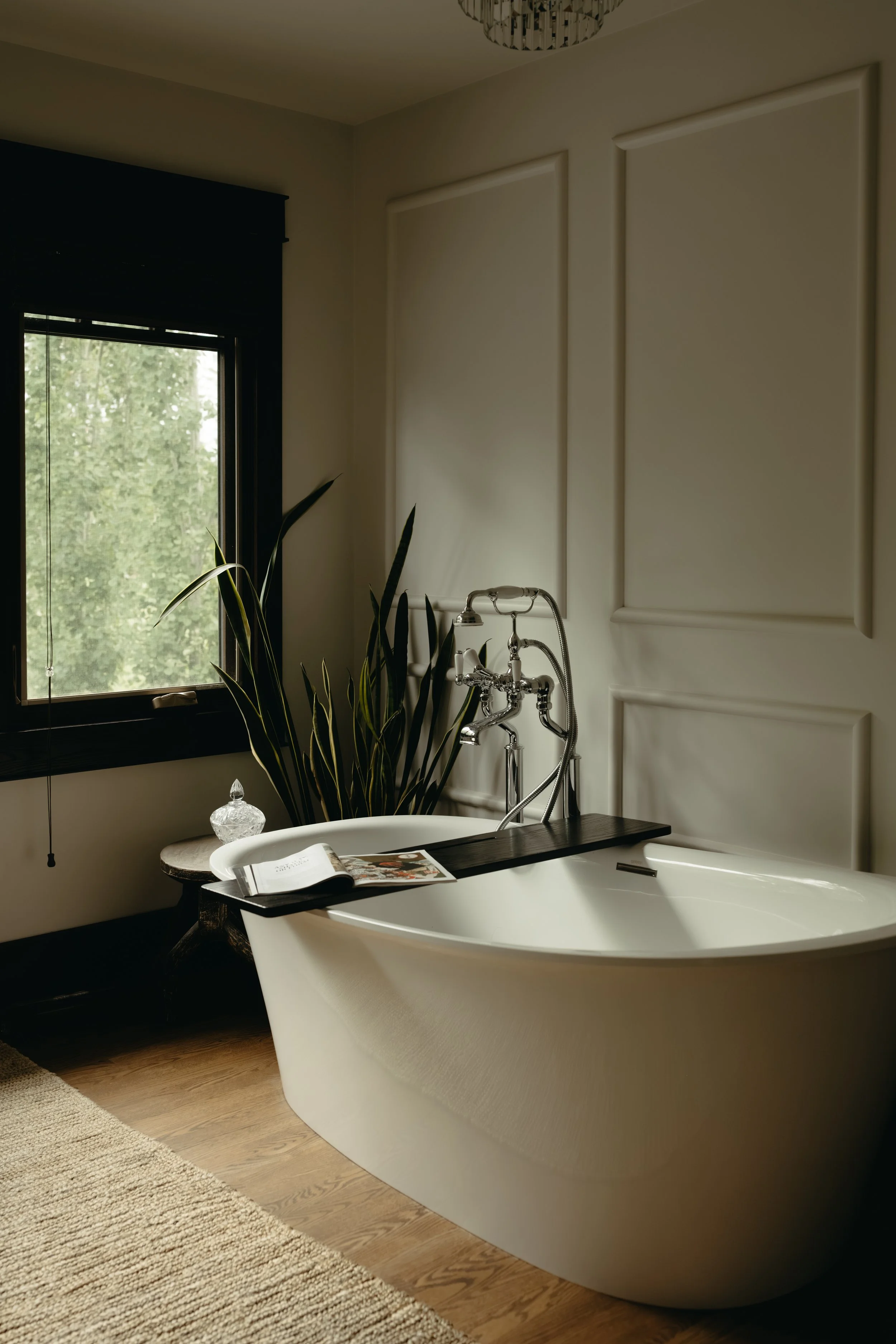 A refined, cozy bathroom featuring a white clawfoot bathtub, a large window overlooking lush greenery, and beautifully curated decor. Every detail is thoughtfully considered to create a luxurious, personalized space designed with intention.