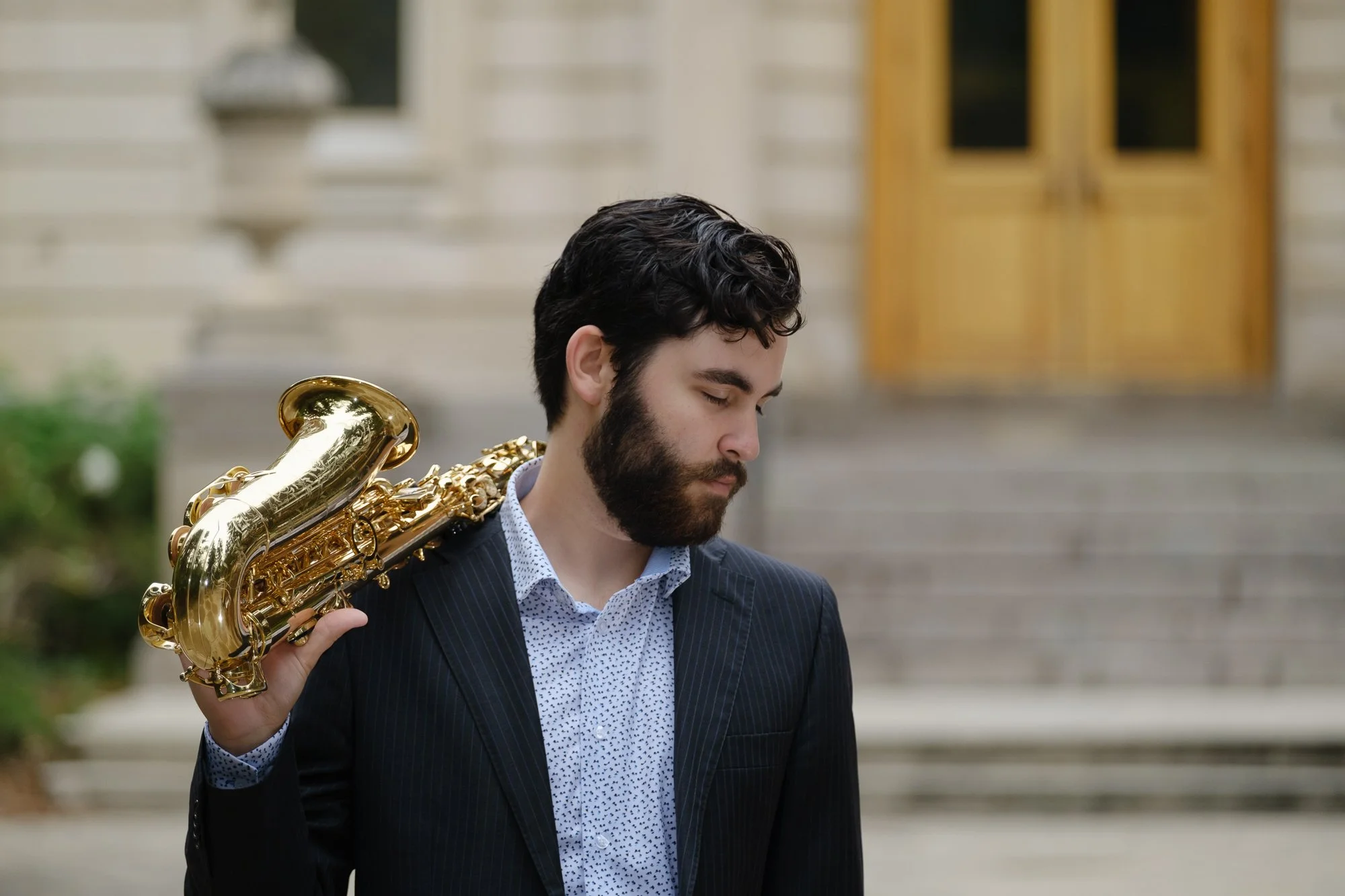 A man with dark curly hair and a beard standing outdoors, holding a shiny gold saxophone on his shoulder, wearing a dark pinstripe suit and a white shirt with small black dots.