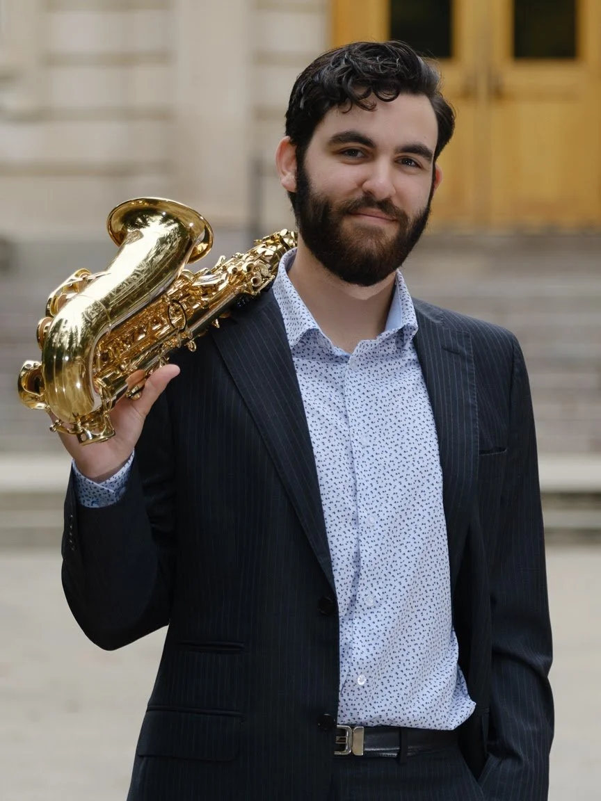 A man in a dark suit holding a gold saxophone over his shoulder, standing in front of stairs and a building with gold-colored doors.