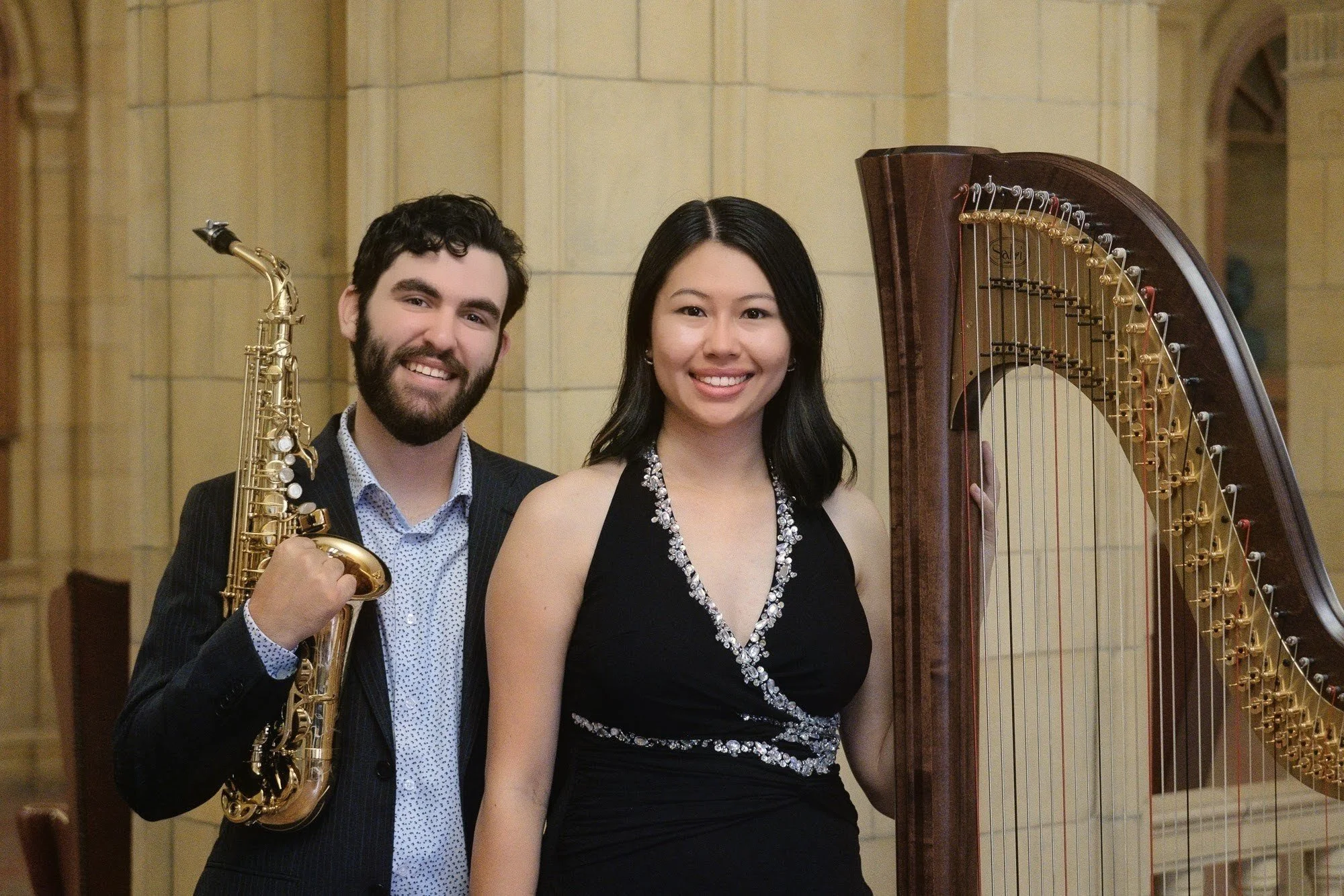Two musicians, a man holding a saxophone and a woman standing near a harp, posing inside a building with beige walls.