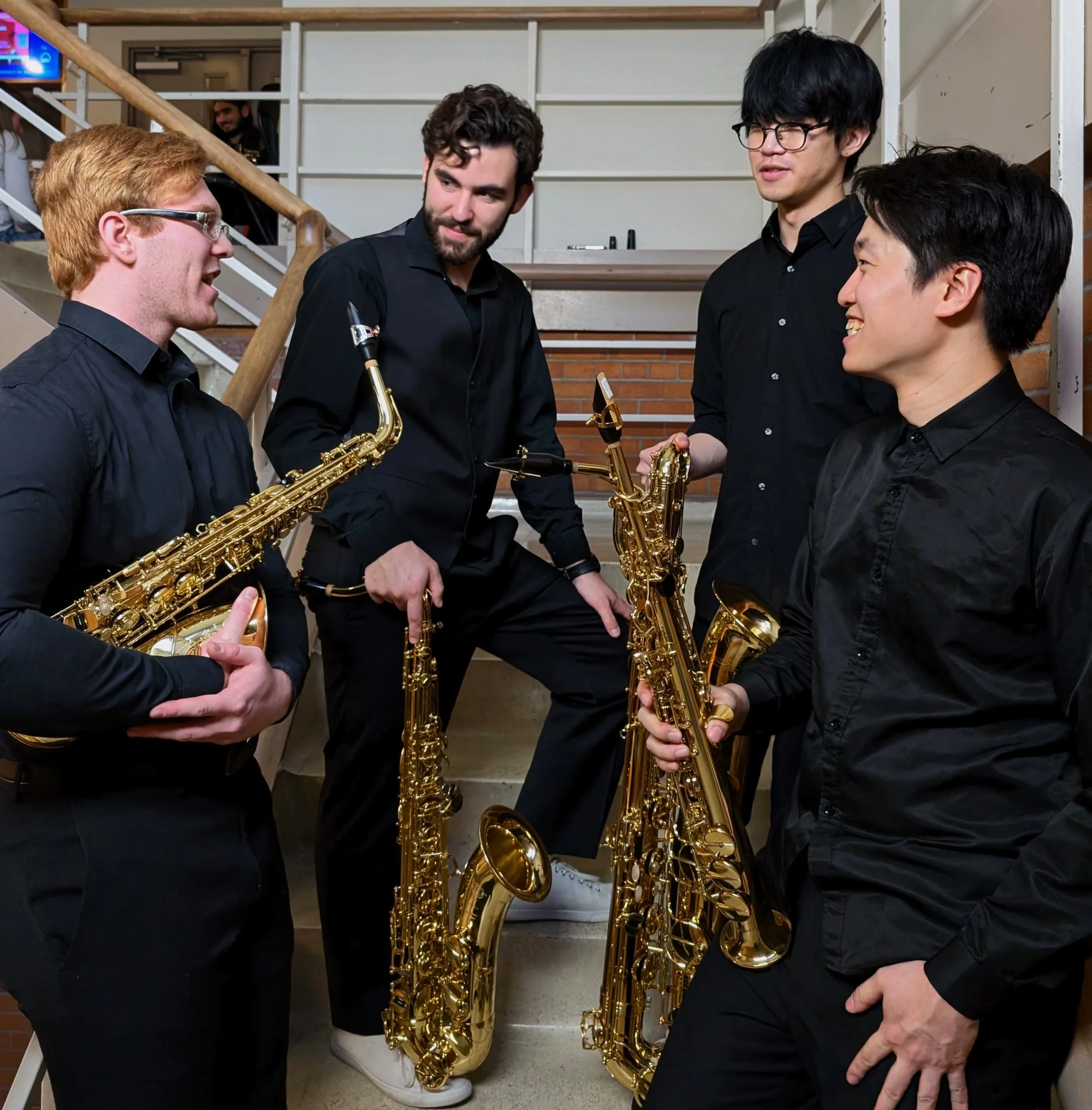 Four men in black shirts holding and displaying gold saxophones, talking and smiling, in an indoor setting with staircase and brick wall in the background.