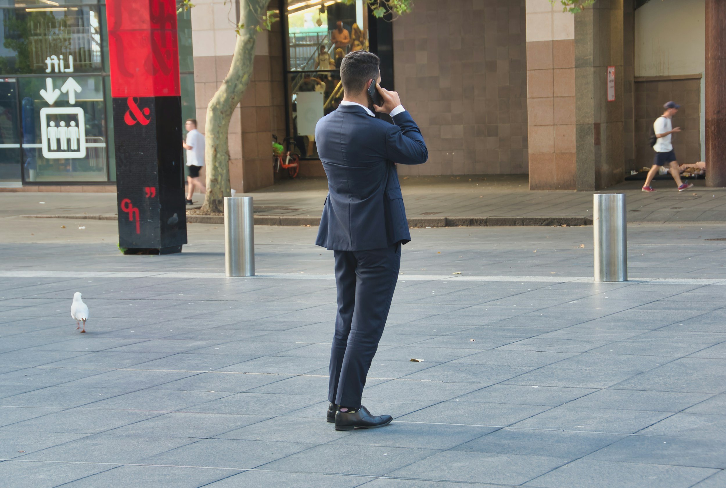 A man in a blue suit standing on a city sidewalk, talking on his cellphone. There are pedestrians, a tree, and a building with large windows in the background.