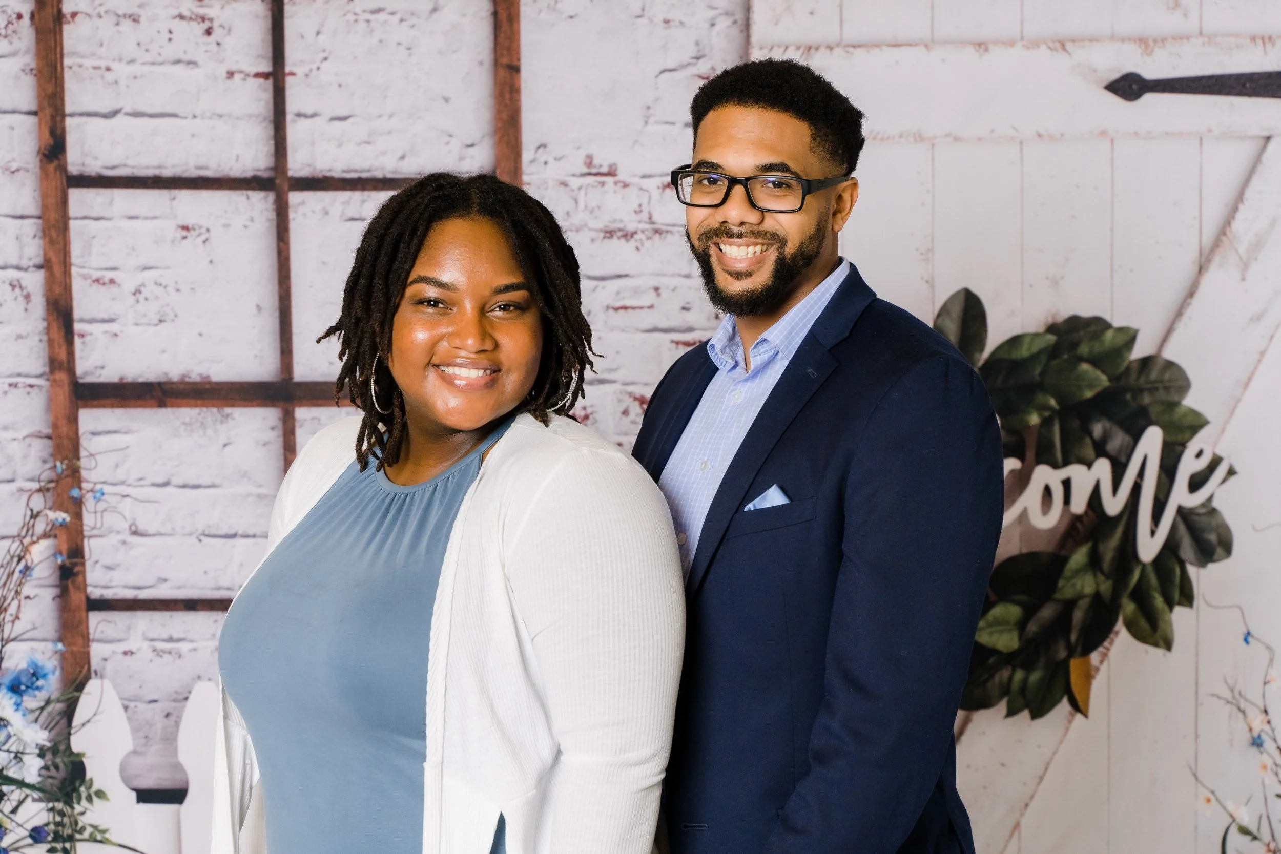 A woman and a man smiling and posing for a photo indoors with a white brick wall background decorated with plants and a wooden frame.