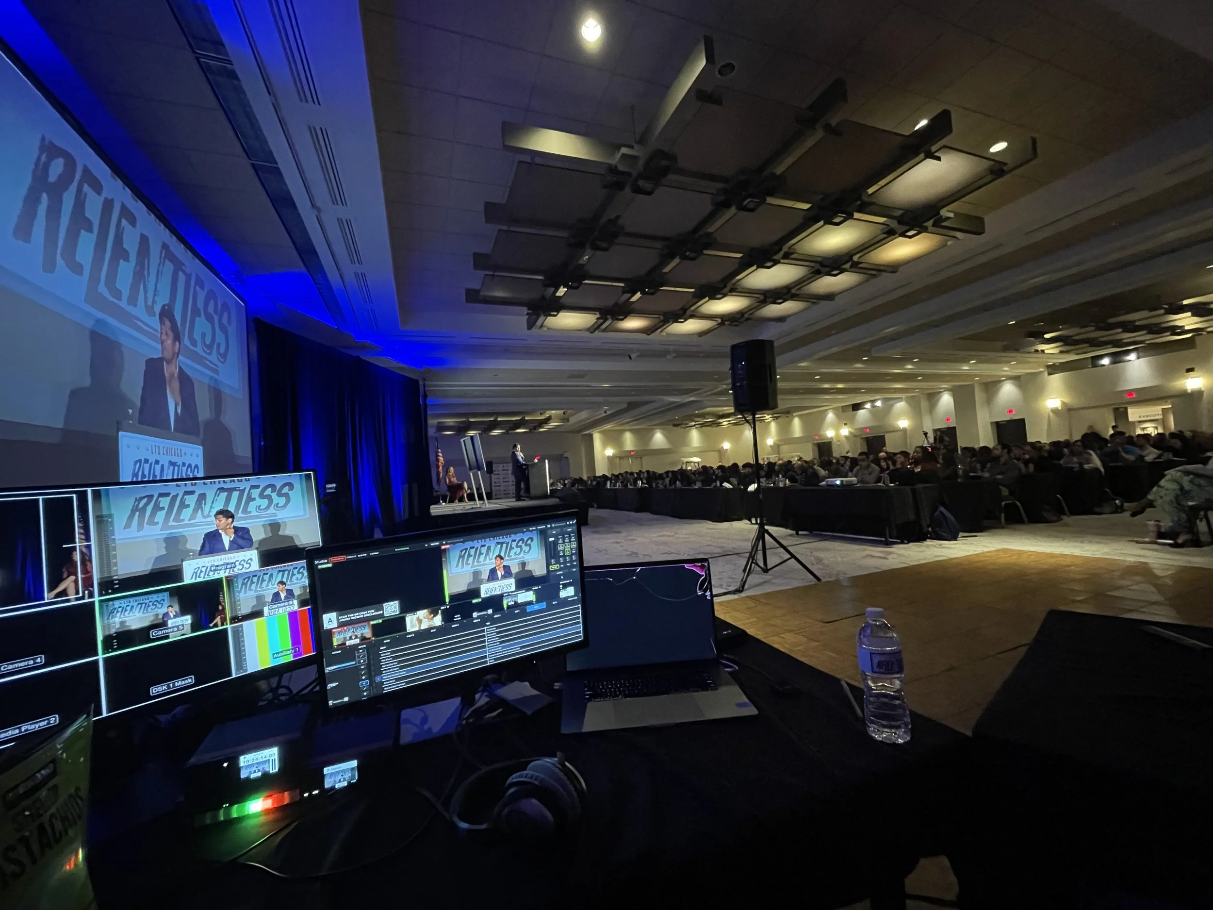 Conference room with large audience, stage, and presentation screen displaying the words 'Relentless' and a person speaking at a podium. Foreground shows a control desk with multiple monitors, sound equipment, and a water bottle.