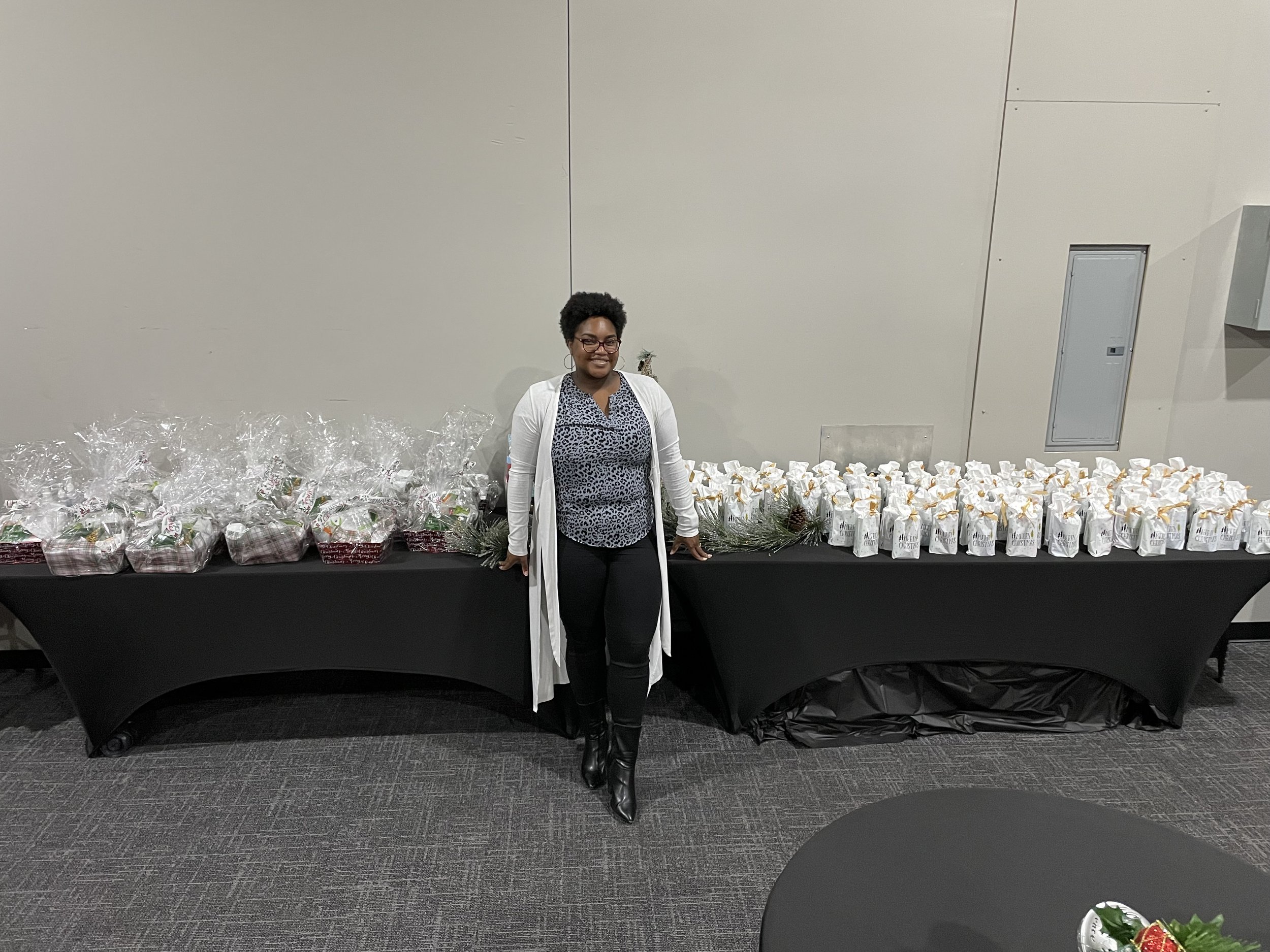 A woman with glasses and a white cardigan standing behind a table filled with white gift bags, decorated with gold bows. The background is a plain white wall with an electrical panel on the right.