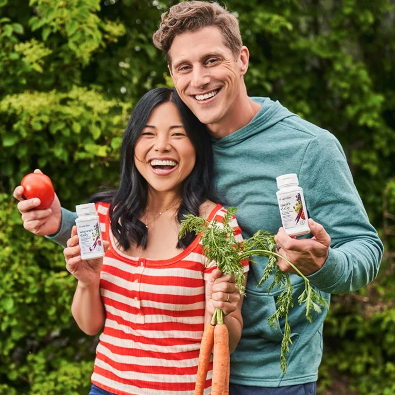 A smiling Asian woman and a smiling Caucasian man outdoors holding vegetables and dietary supplement bottles.