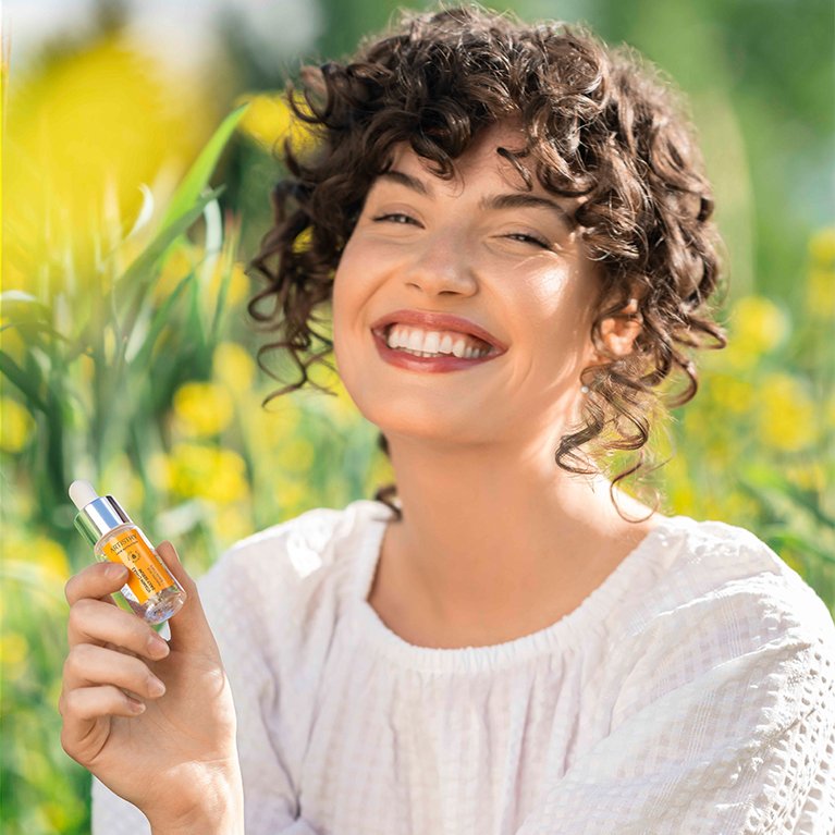 A woman with curly hair smiling outdoors, holding a small bottle of skincare serum, standing in a field of yellow flowers.