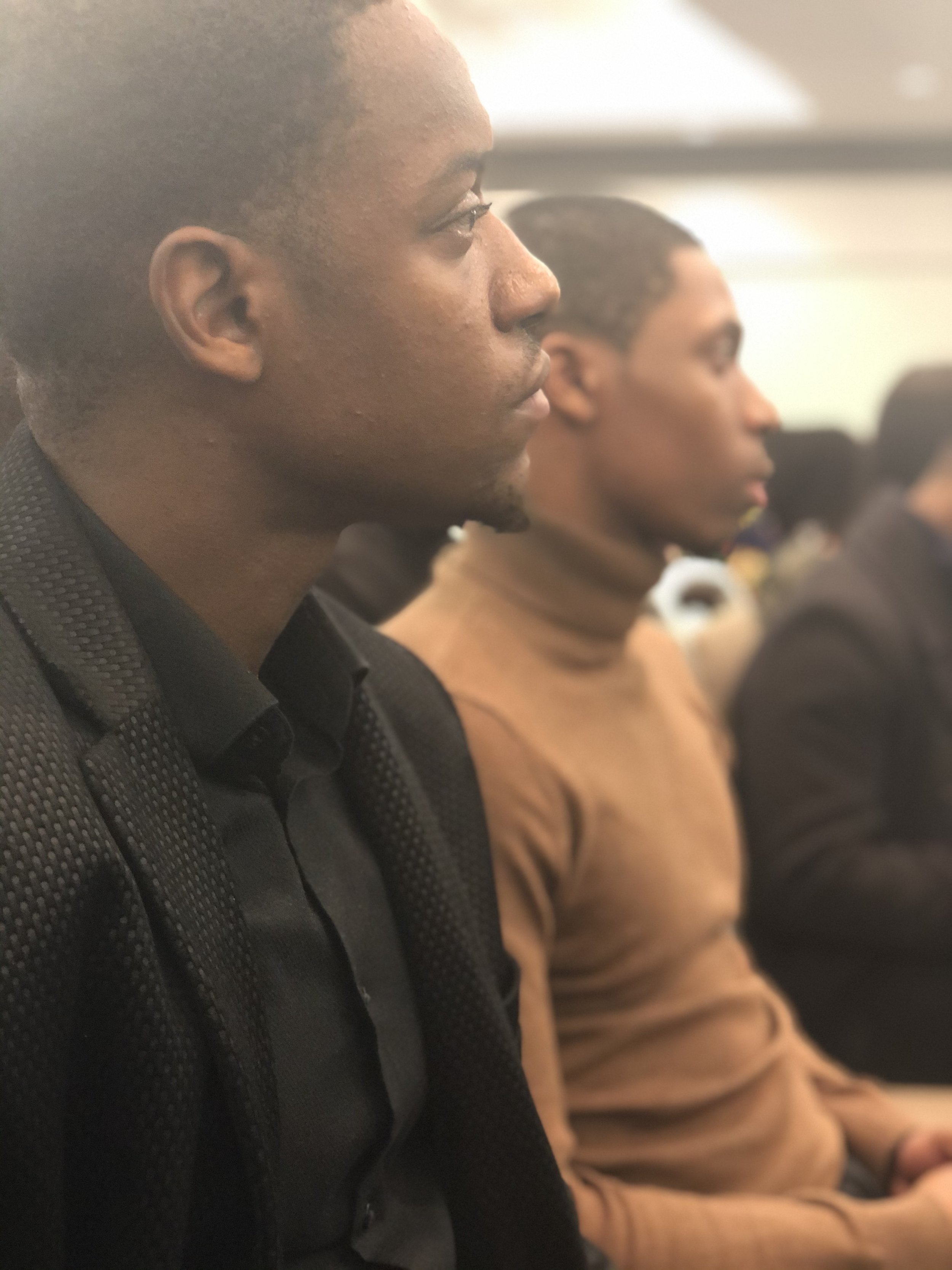 Side view of two people sitting in a row, one man in focus wearing a black suit and the woman in the background wearing a brown jacket, both appearing attentive.