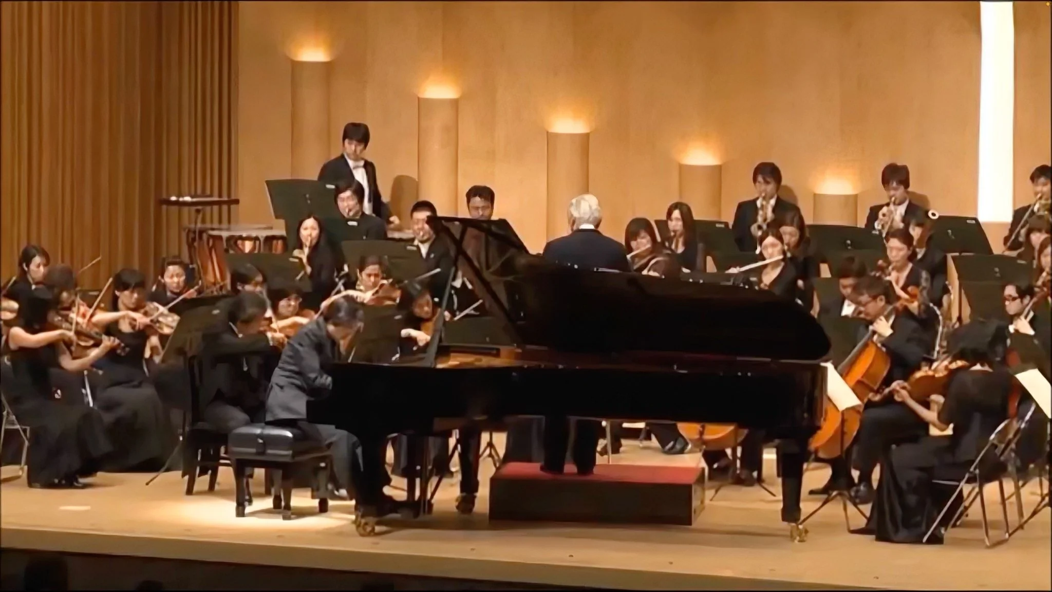 オーケストラのコンサート風景。ピアノ、バイオリン、フルートなどの楽器を演奏する音楽家と指揮者が舞台にいる。