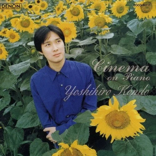男の人がひまわりの花畑に立っている写真で、青いシャツを着ている。