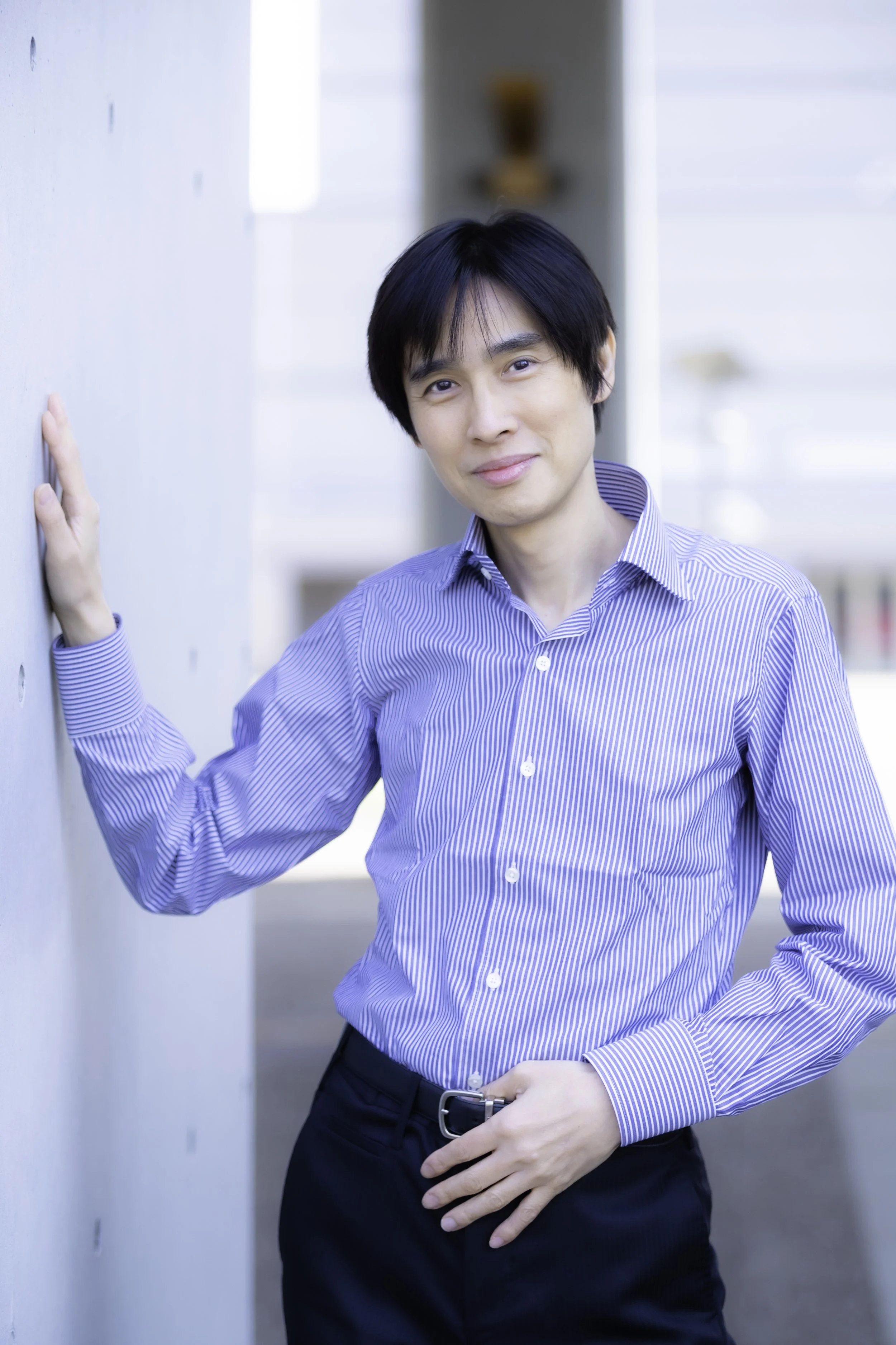 ビジネスマンの男性が白と青のストライプシャツを着て、壁に寄りかかりながら微笑んでいる。」}】}