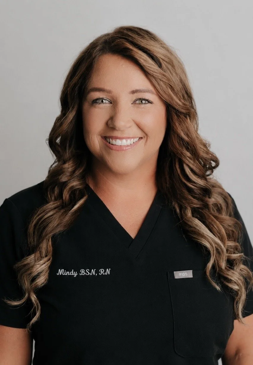 A smiling woman with long, wavy brown hair wearing a black medical scrubs top with her name and credentials, 'Mindy BSN, RN,' embroidered on it.