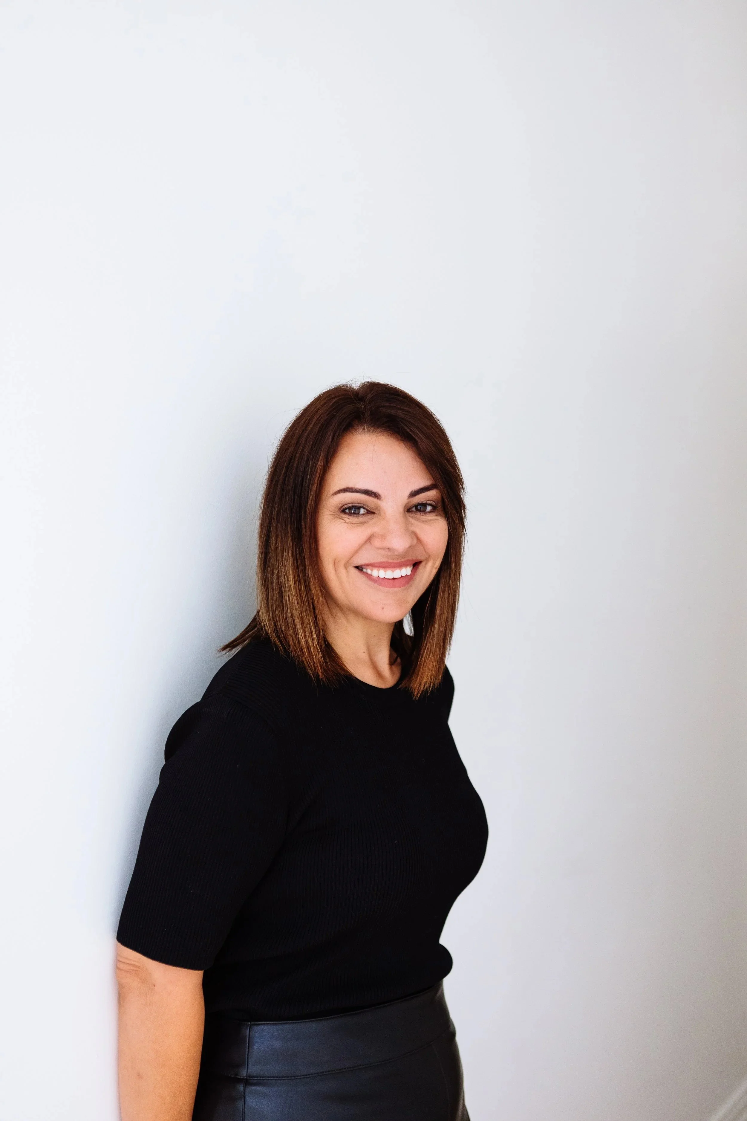 A woman with shoulder-length brown hair smiling, wearing a black top and black skirt, standing against a plain white wall.