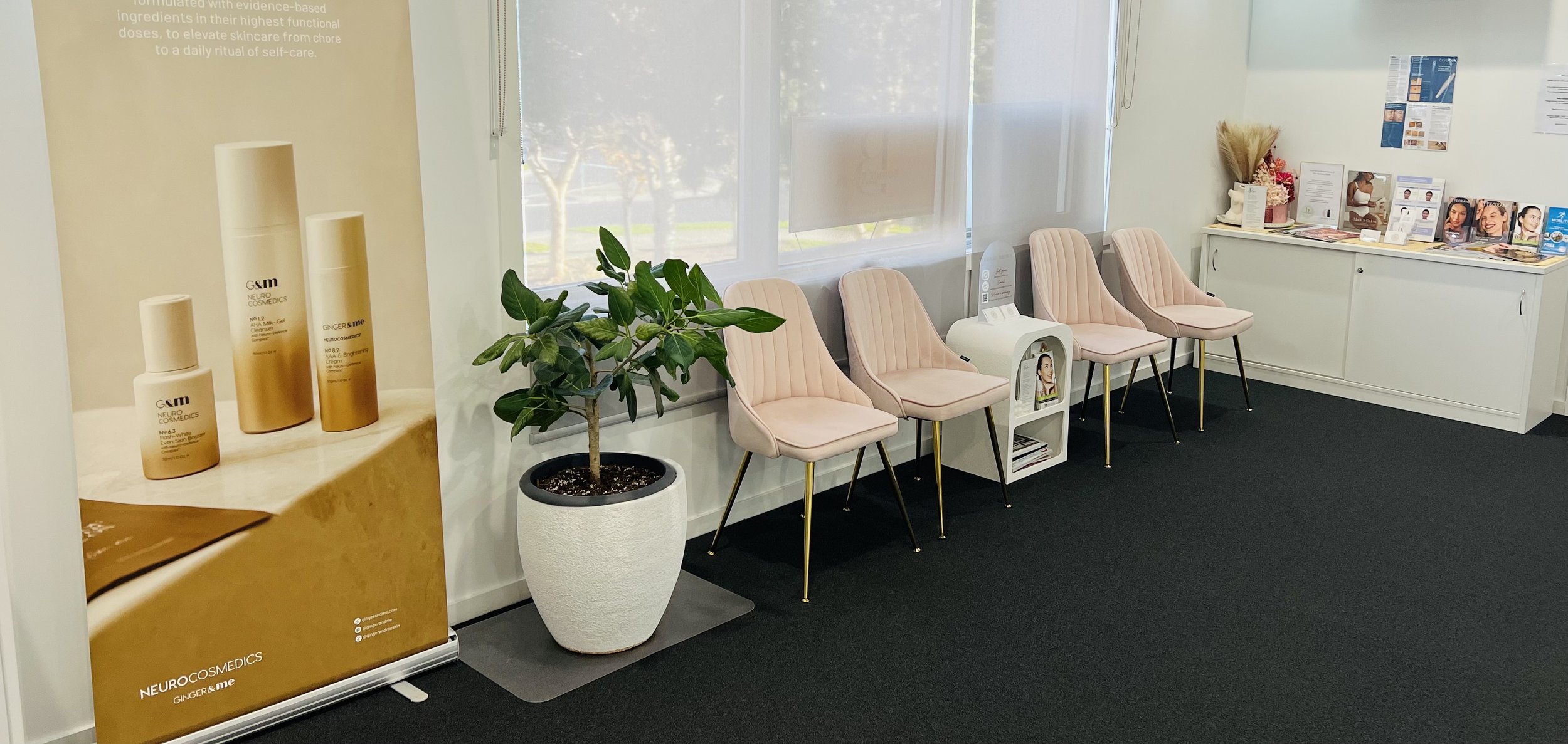 A waiting room with four pink upholstered chairs with gold legs, a potted plant, and a white cabinet with pamphlets and brochures. There are windows with blinds and a large poster display for Neurocosmetics products, including bottles of skincare.