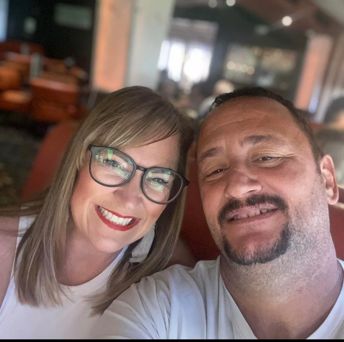A smiling woman with glasses and red lipstick taking a selfie with a smiling man with facial hair in a restaurant or cafe.