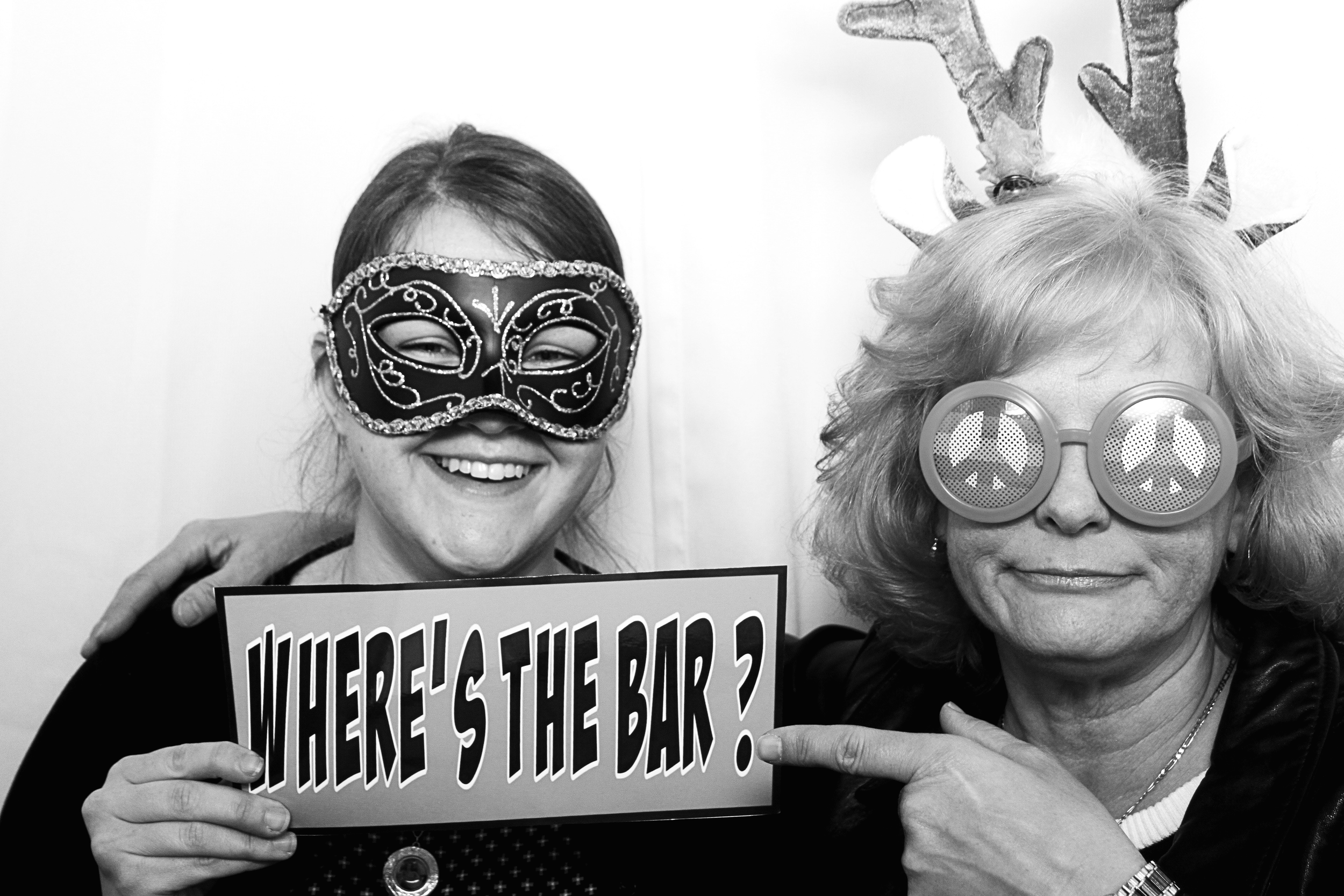 Two women in a photo booth wearing costumes and masks with peace sign glasses, holding a sign that says 'WHERE'S THE BAR?'.