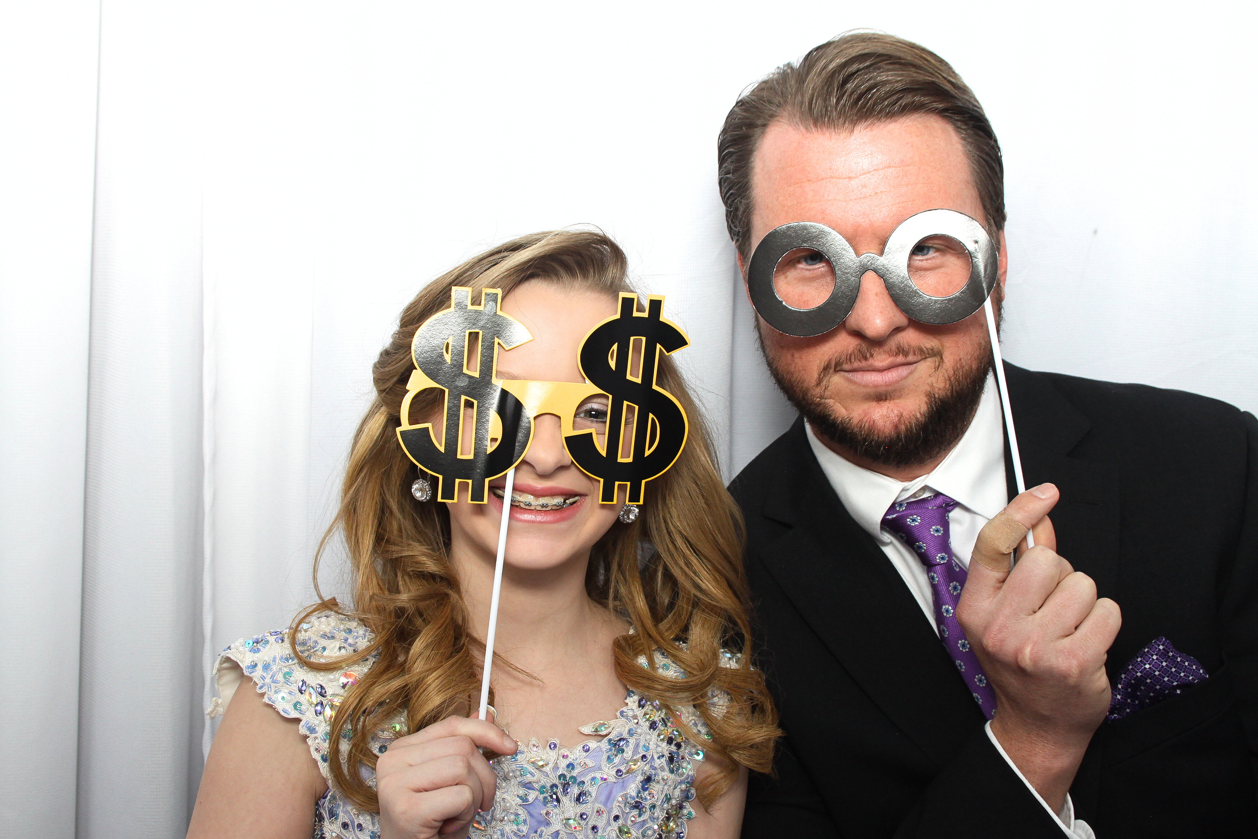 A young girl and a man in a photo booth wearing novelty glasses with dollar signs and the man with glasses with large circles are smiling and holding photo booth props against a white background.