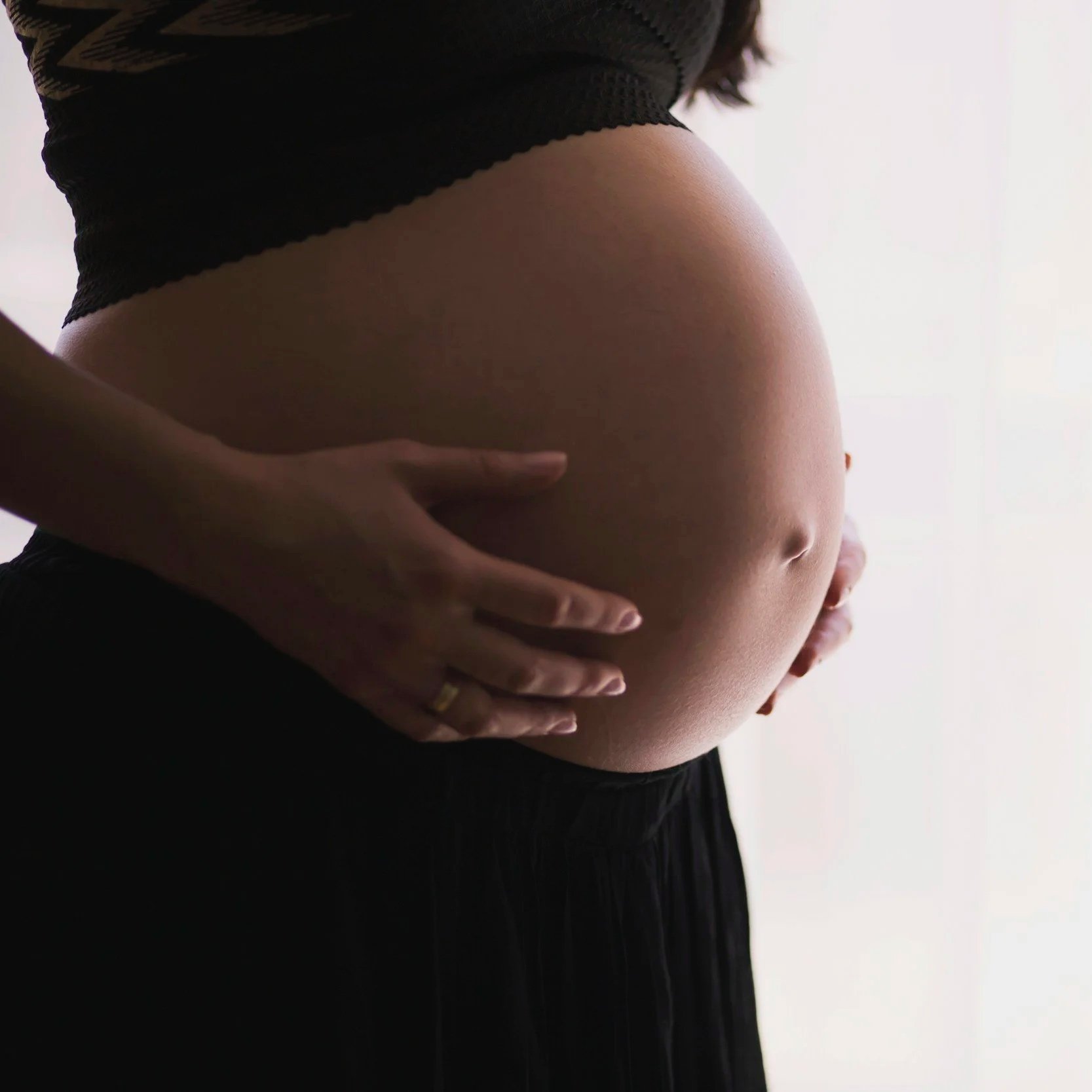 Close-up of a pregnant woman's belly being gently held by her hands, with the woman wearing a black top and skirt.