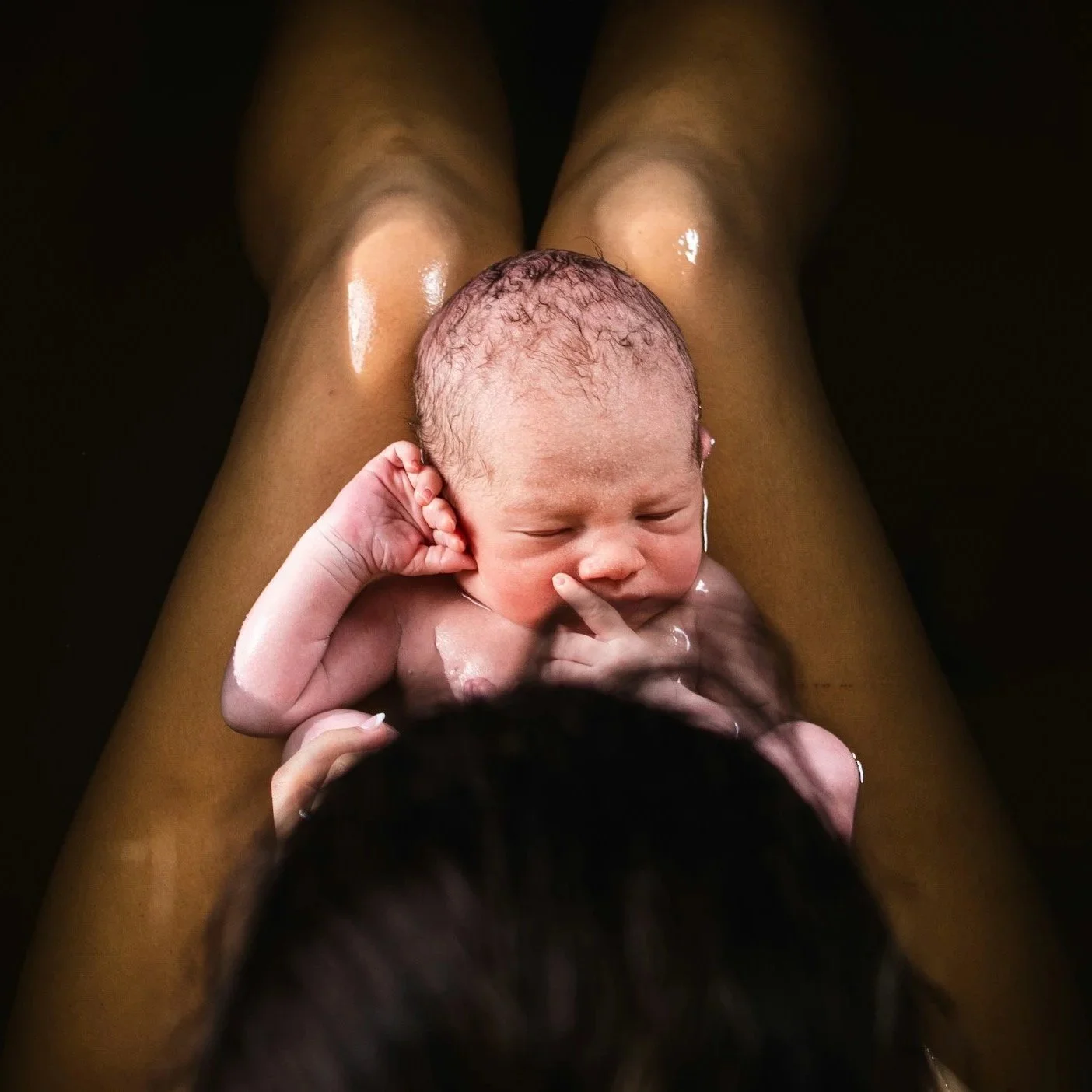A newborn baby lying on someone's legs, with water on their skin and hair, sleeping peacefully with one hand near the face and the other resting on the arm.