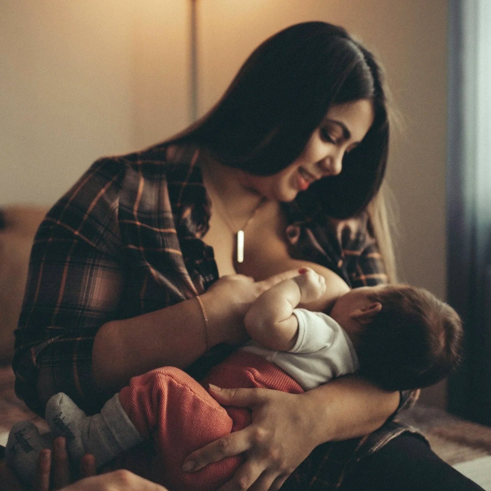 A woman with long dark hair smiling while leaning over a child, possibly giving a hug or comforting them, indoors with warm lighting.