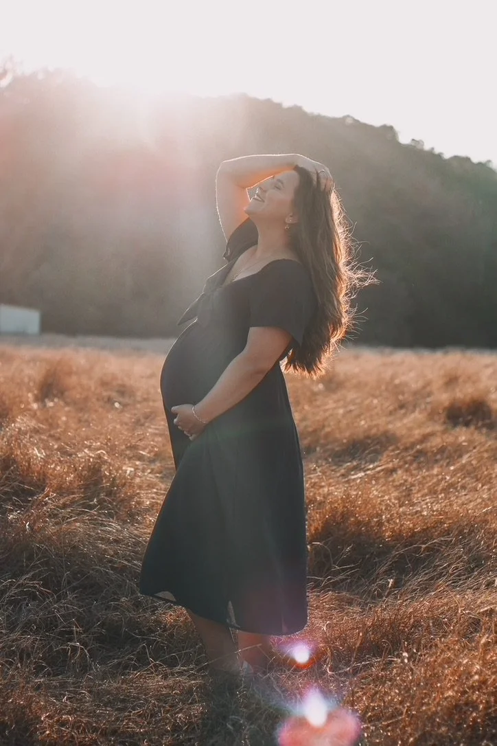 A pregnant woman in a black dress standing in a field with tall grass at sunset, smiling and touching her hair.