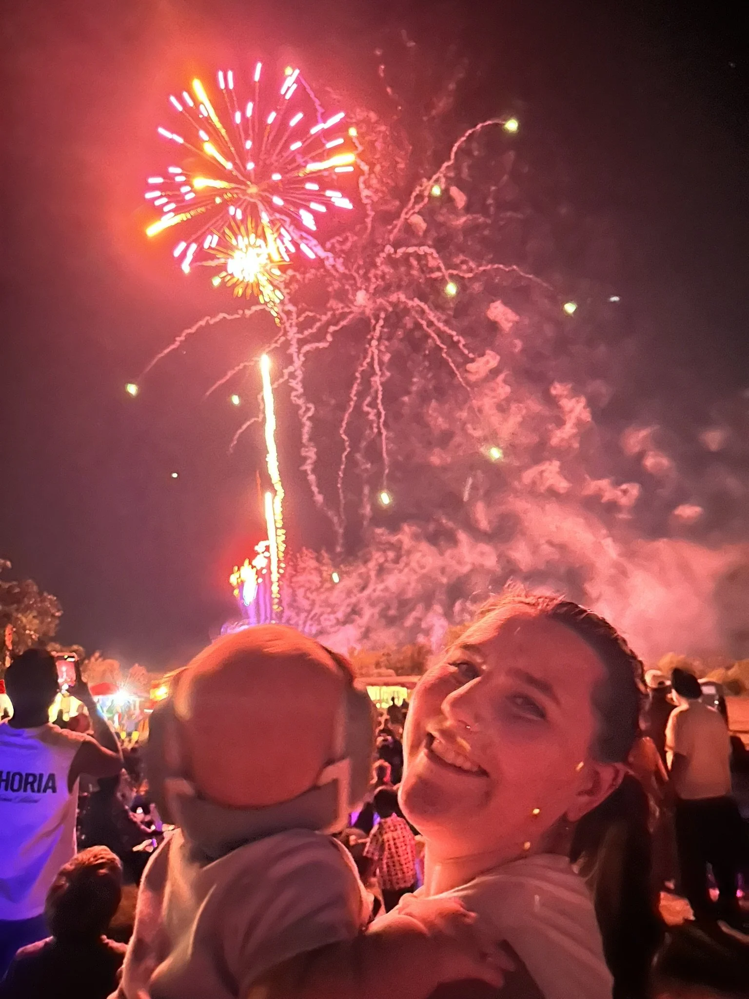 People watching fireworks display at night, with a smiling woman in the foreground and others capturing the event on their phones.