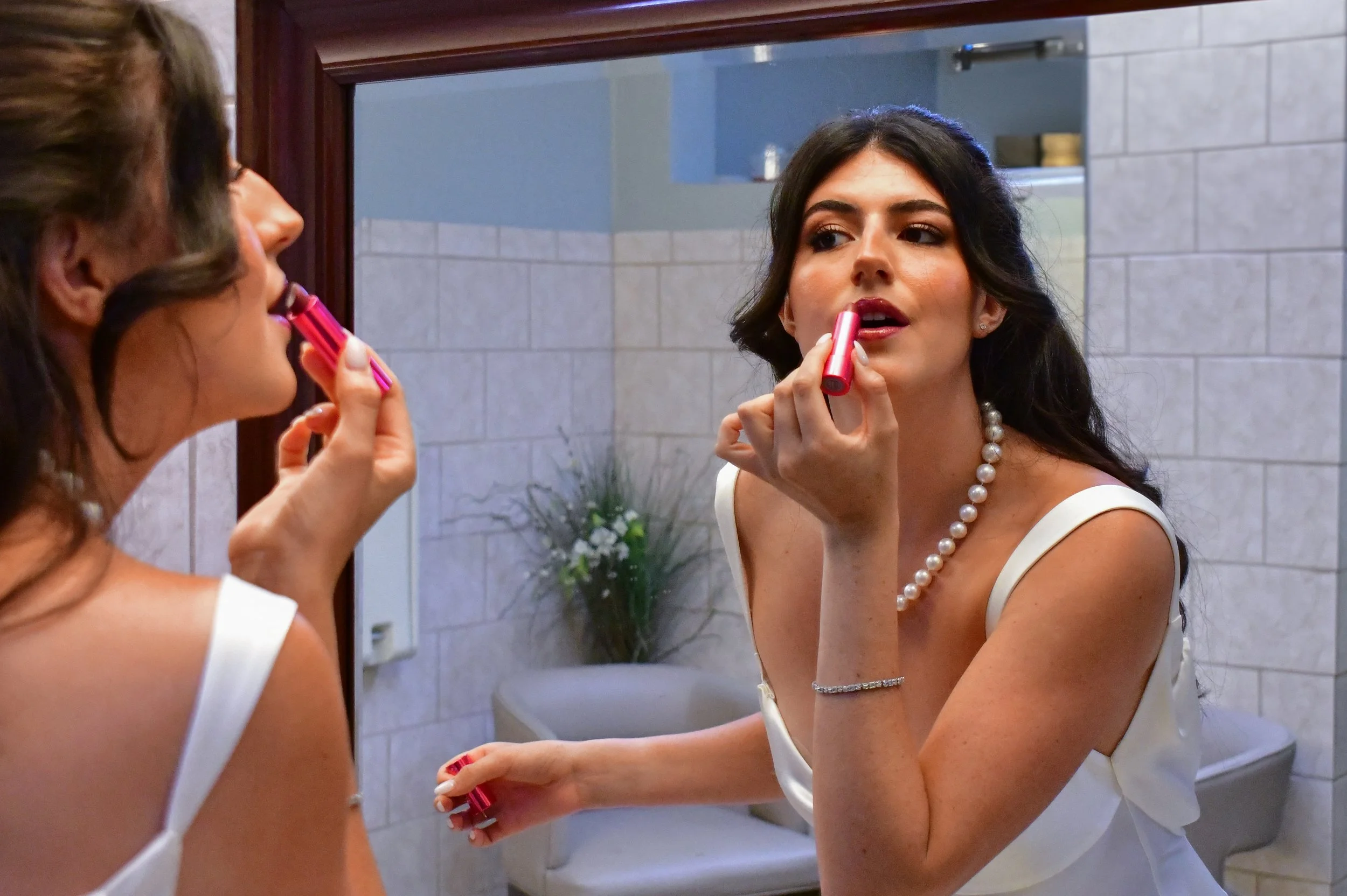 Woman applying lipstick while looking in a mirror in a room with tiled walls and a plant in the background.