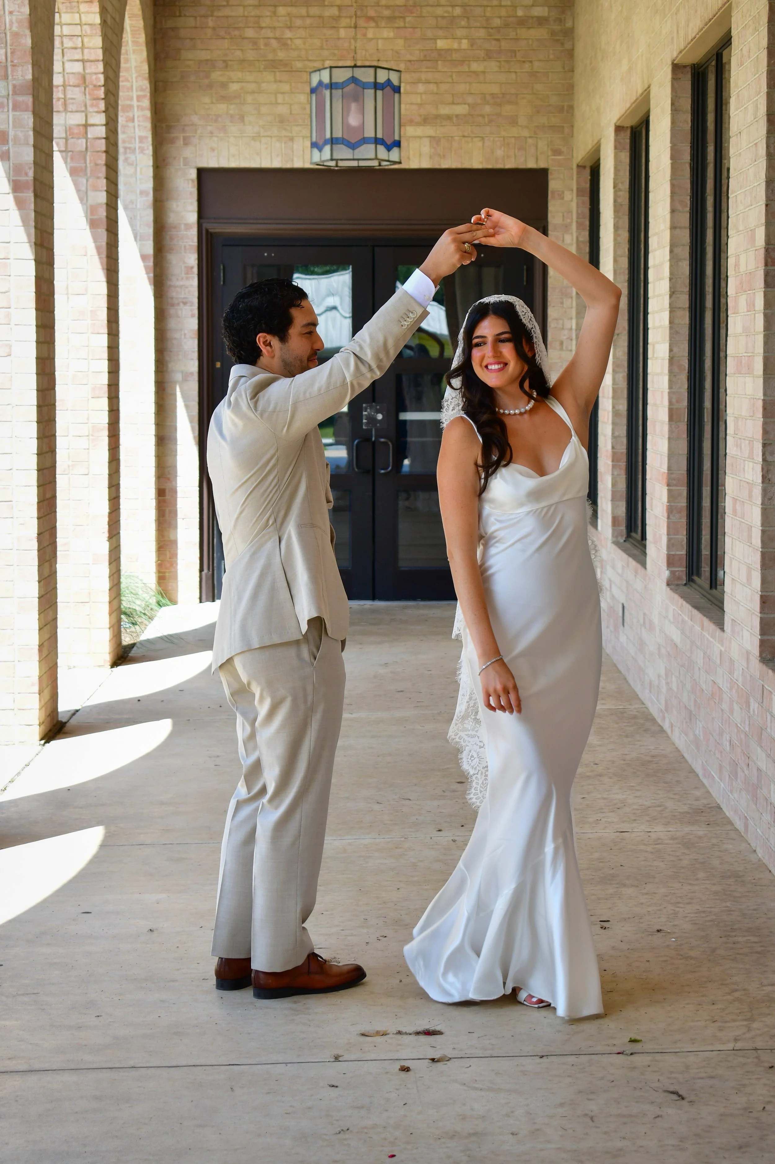 A couple dressed in wedding attire dancing. The man is wearing a beige suit and brown shoes, and the woman is in a white wedding gown with a veil and pearl necklace.