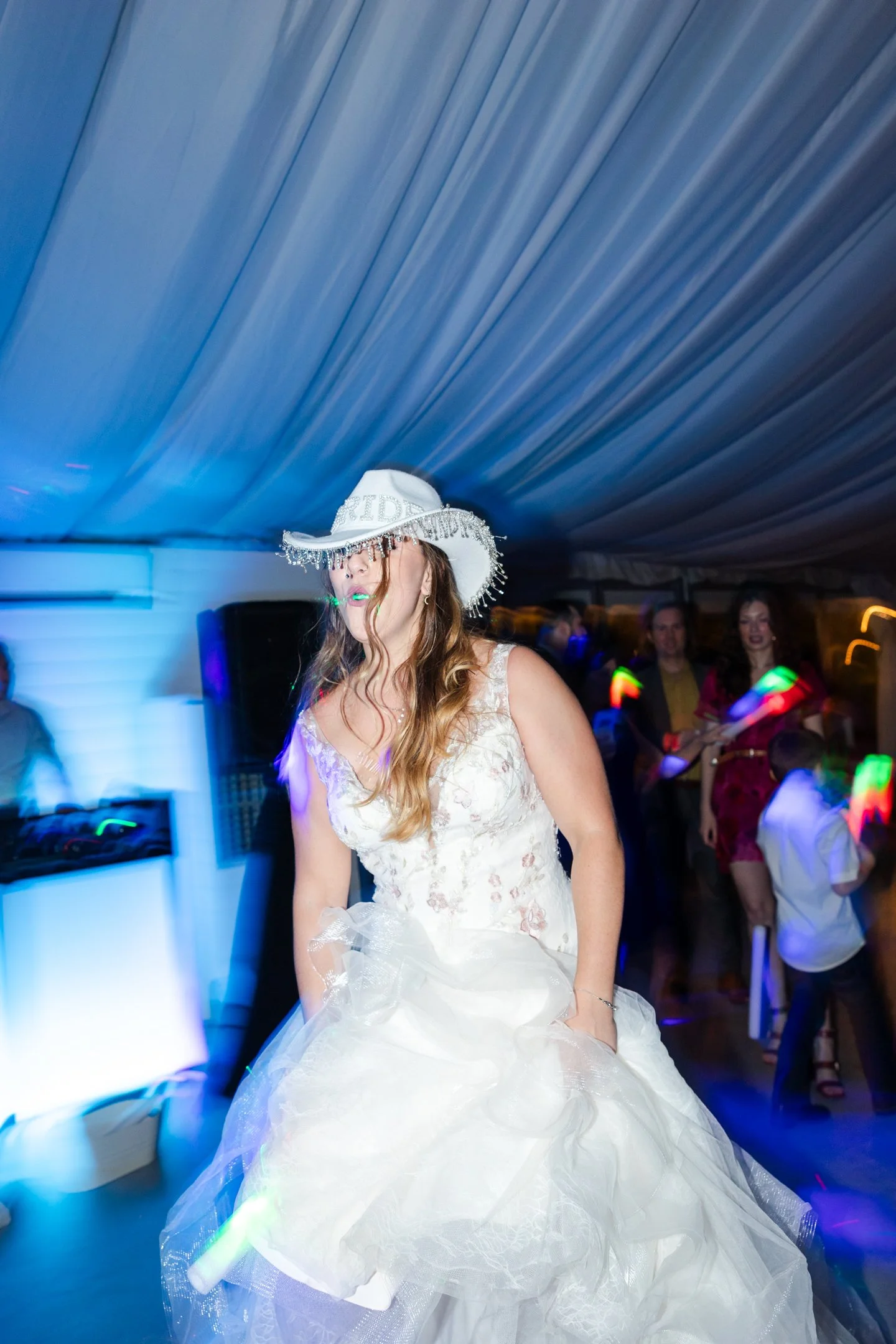 Julia, photographer and owner of JEM Media, in a wedding dress and a white cowboy hat with silver embellishments, dancing at a party with colorful lights and other guests in the background.