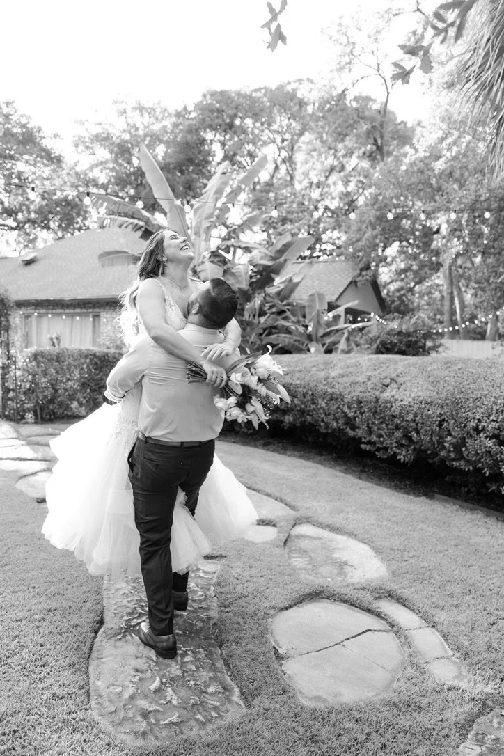 A groom lifting a bride in a garden setting with string lights overhead, trees, and a house in the background