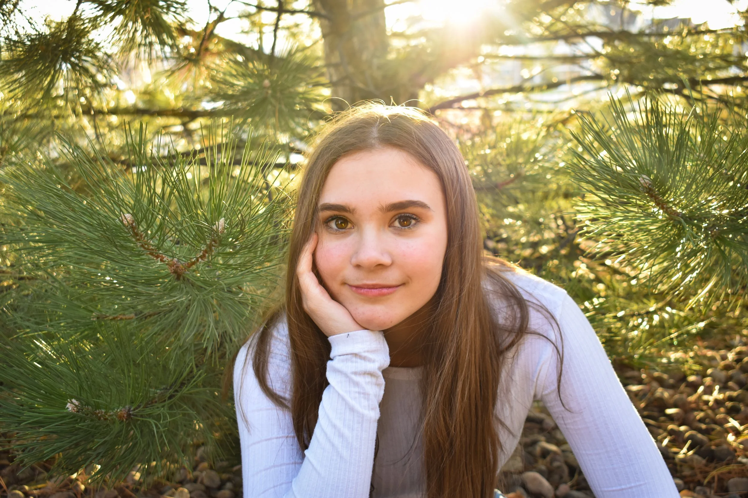 A young woman with long brown hair and light skin lying on the ground among pine tree branches, with sunlight shining through the trees behind her.