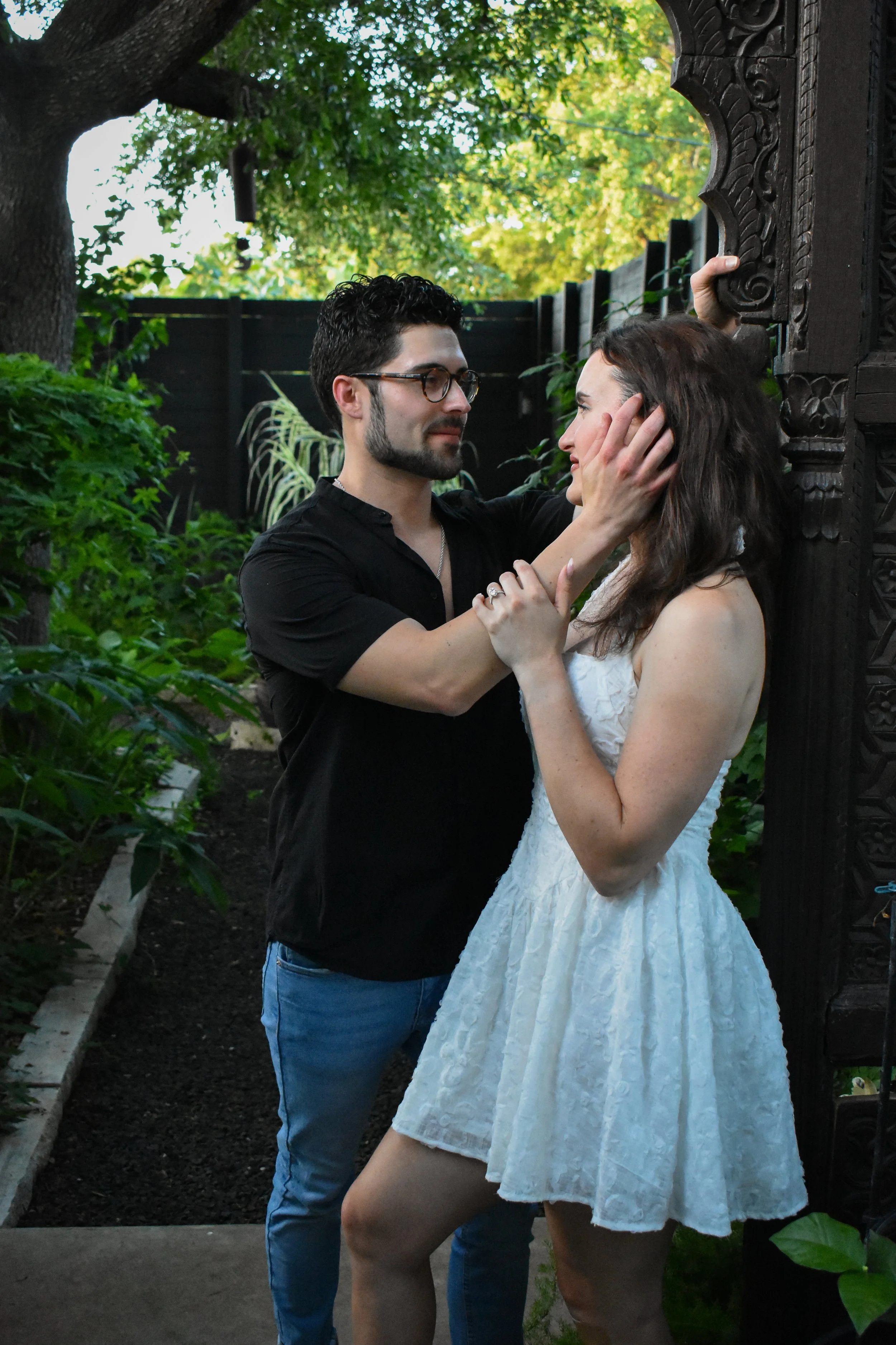 A man and woman in a garden pose closely together, with the man touching the woman's face and the woman holding his arm. The man is wearing glasses and a black shirt, while the woman is in a white dress.