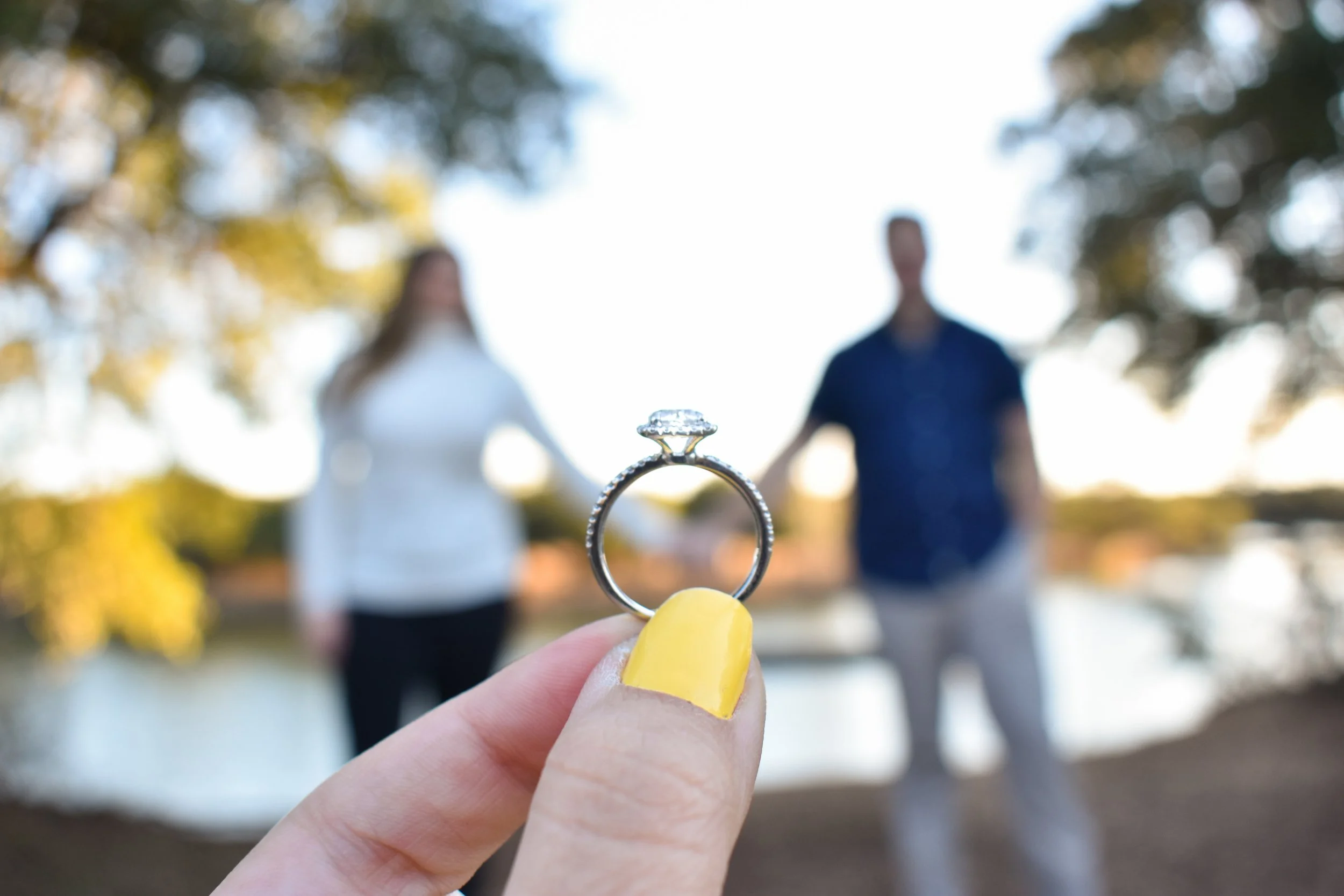 A close-up of a hand with yellow painted nails holding a ring between the thumb and index finger. In the background, a blurred couple stands outdoors under trees, holding hands and facing each other in a park or garden setting during daytime.