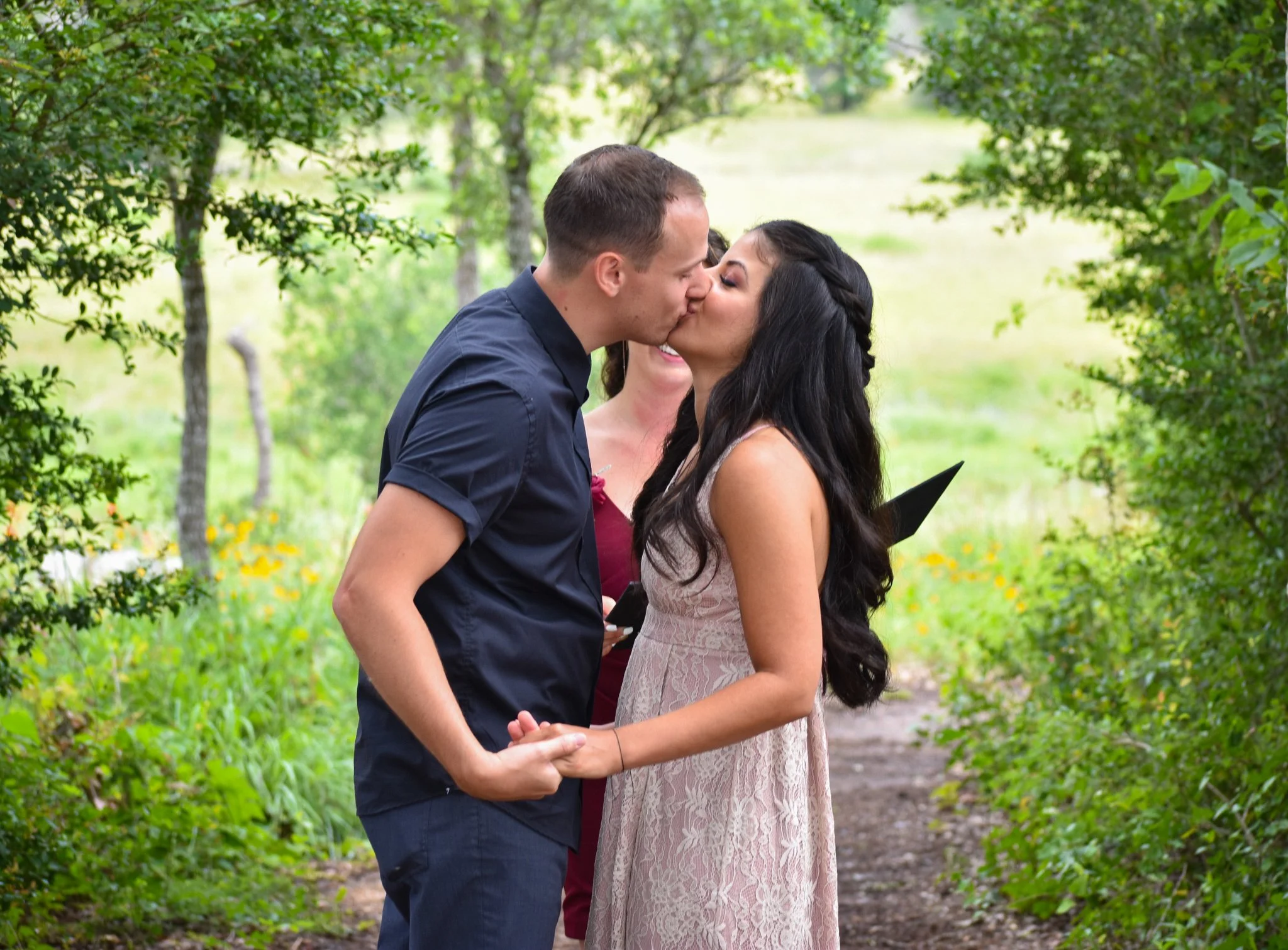 A couple sharing a kiss while holding hands outdoors in a lush, green park.