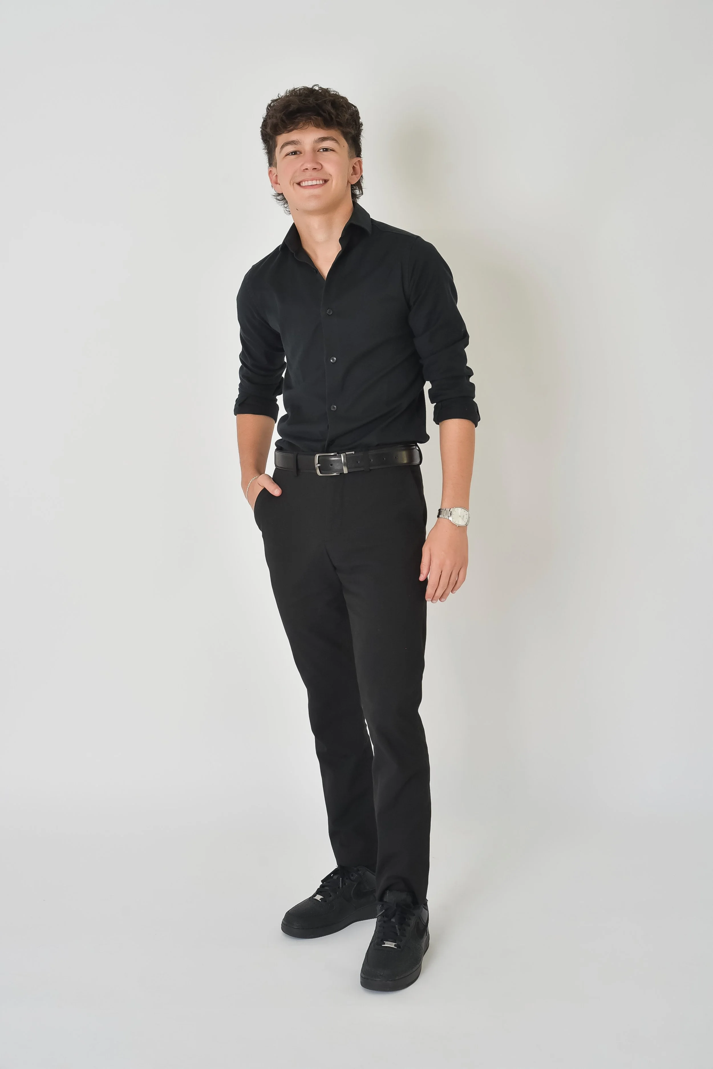 Young man smiling, wearing black shirt, black pants, black sneakers, standing against a plain white background.