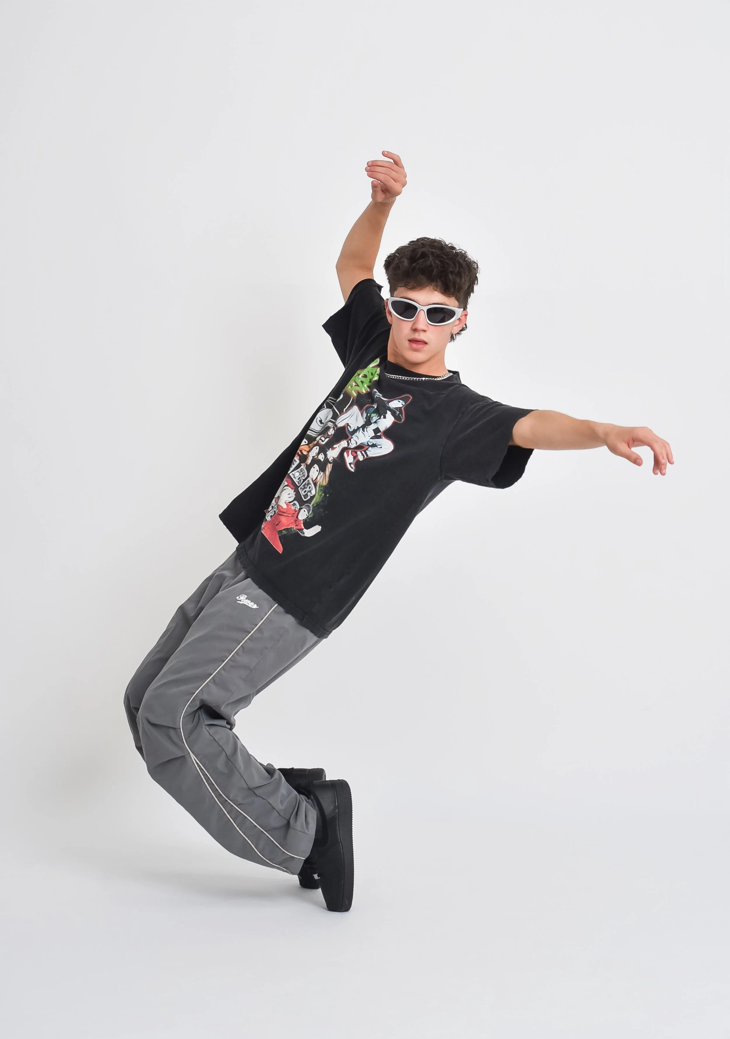 Young man wearing black graphic T-shirt, gray sweatpants, black boots, white sunglasses, and a silver chain necklace poses standing on one leg with arms outstretched against a plain white background.