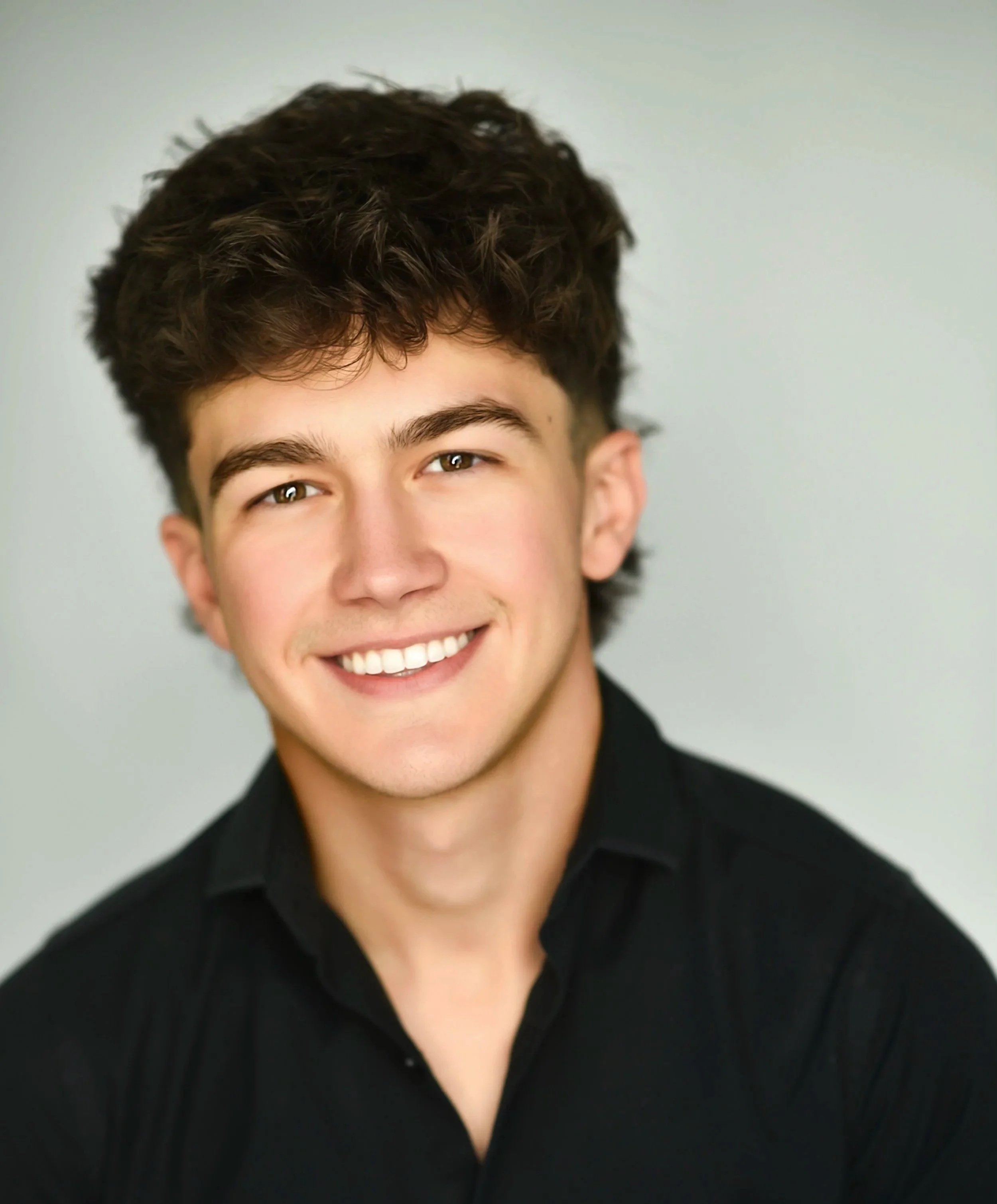 Close-up of a smiling young man with dark, curly hair, wearing a black shirt.