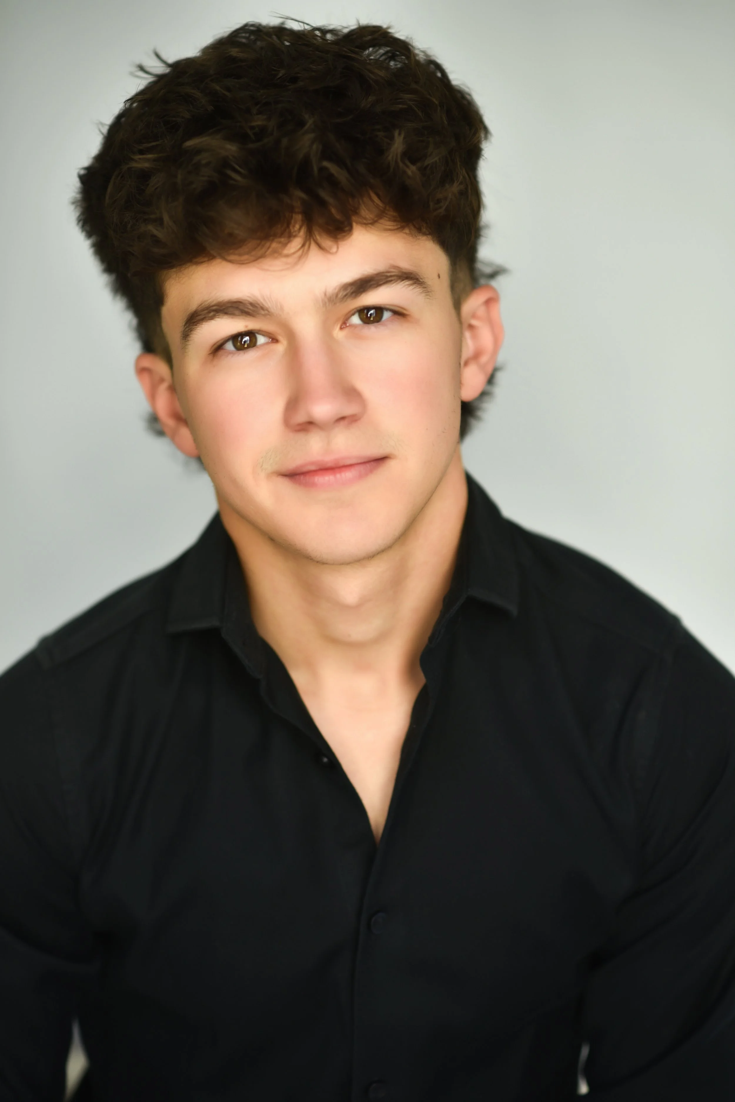 Portrait of a young man with brown curly hair and hazel eyes, wearing a black shirt against a light gray background.